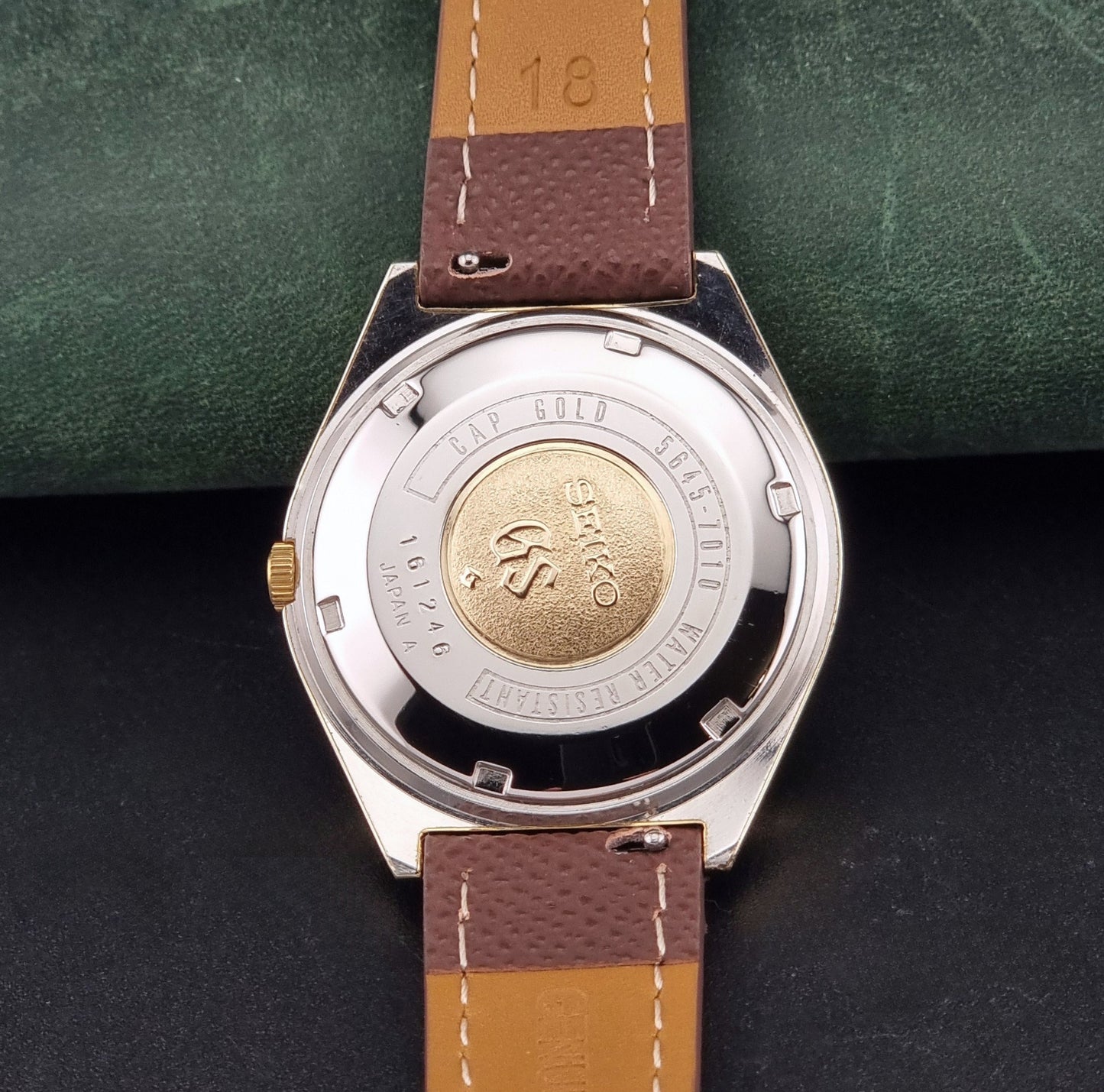 Vintage Grand Seiko Wristwatch 5645-7010 with a gold-tone case, cream dial, and brown textured leather strap, photographed on a dark gray background case back view