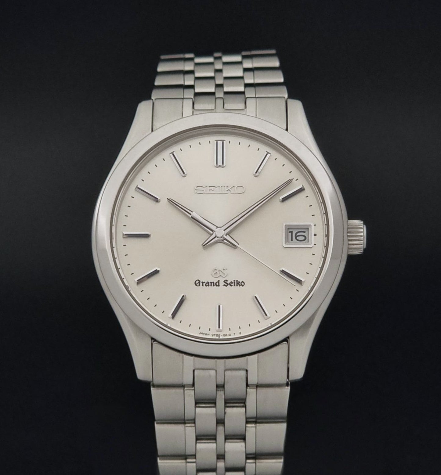 A Grand Seiko SBGV001 quartz watch and link bracelet with a cream-colored dial, and a date window, shown against a dark background.