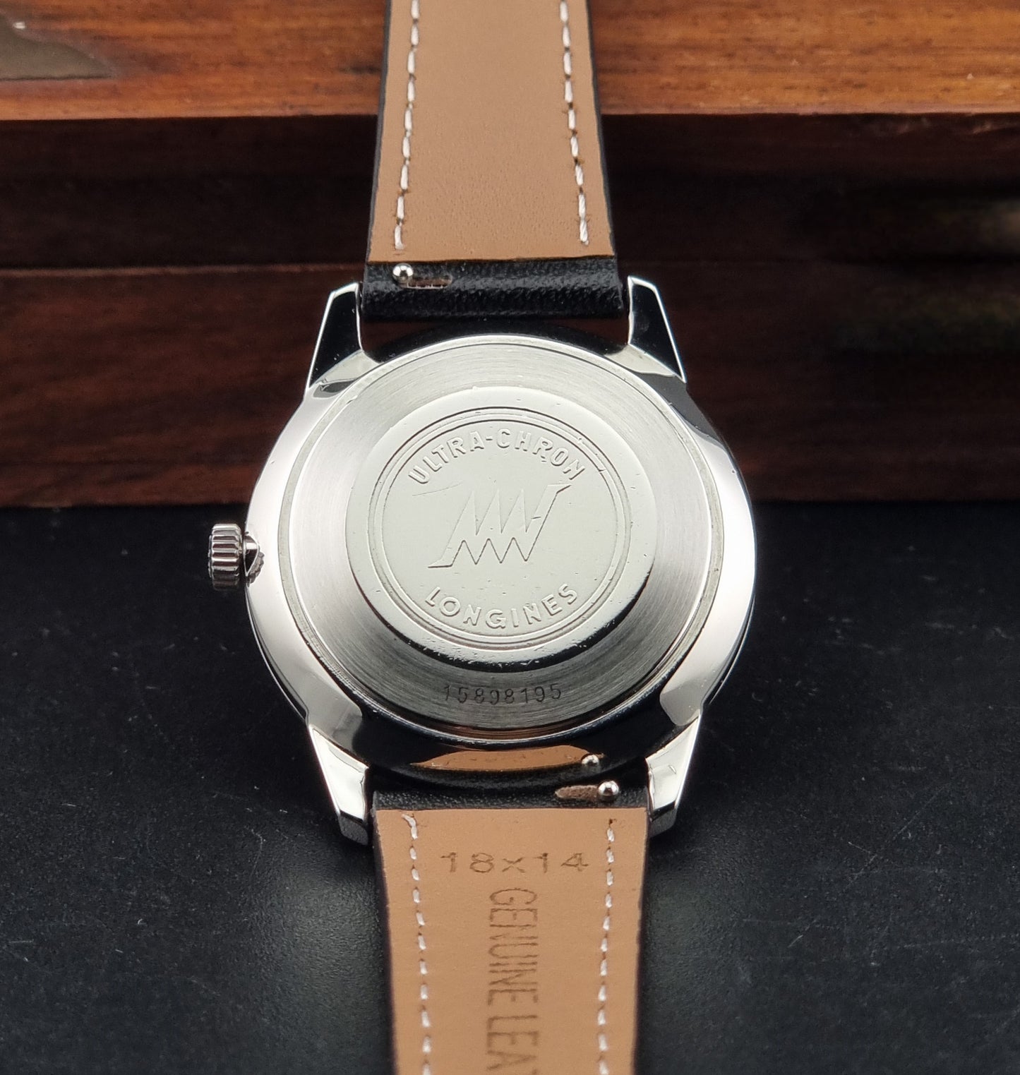 Close-up of the back of a Longines wristwatch with a silver metal case and a tan leather strap, resting on a dark surface with a wooden background.