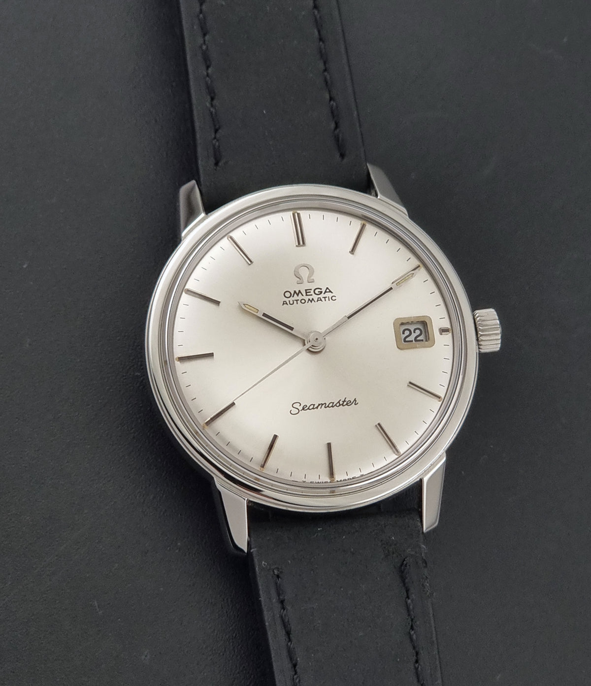 An Omega Seamaster wristwatch with a silver-toned stainless steel case, a white dial, and a black leather strap, shown on a dark textured background.