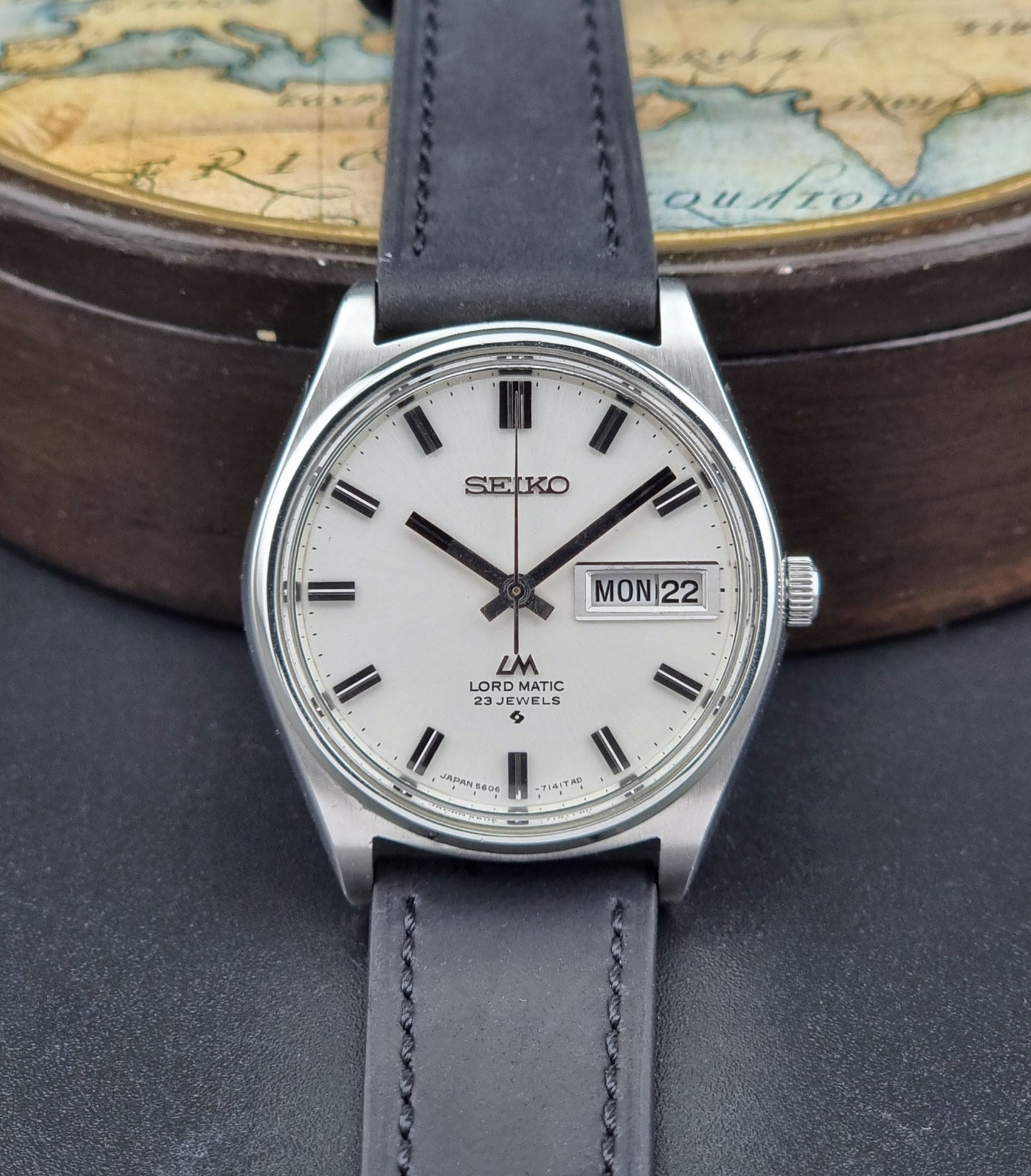 Vintage Seiko Lord Matic watch with gray leather strap on a dark surface with a world map in the background