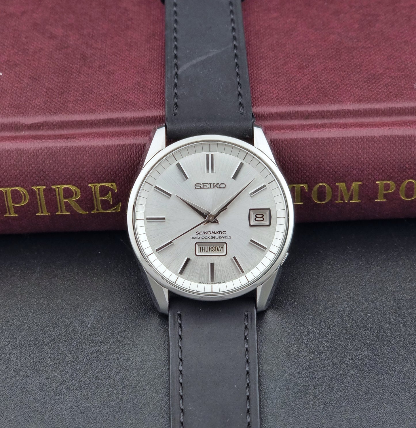 Seiko Seikomatic 6206-8040 Wristwatch with a silver dial and black leather strap on a dark surface with a book in the background.
