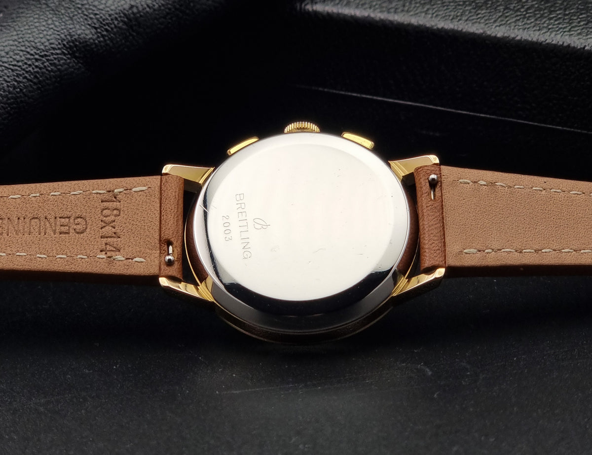 Close-up of the back of aVintage Breitling Top Time 2003 Chronograph watch with a tan leather strap and gold-tone case, showing the Breitling logo and model text on the polished metal case against a dark background.