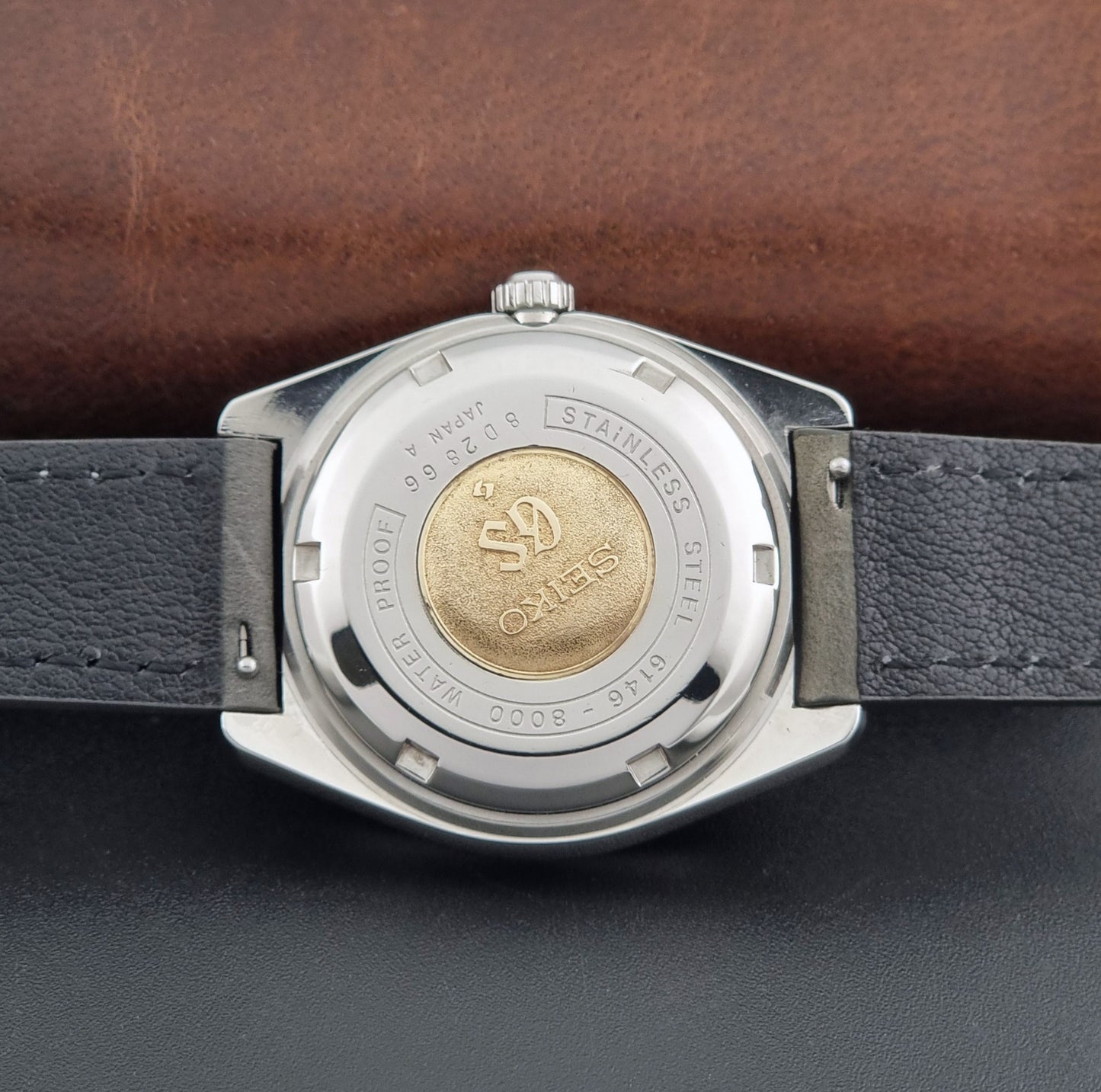 Vintage Grand Seiko Wristwatch 6146-8000 with gray strap and silver case on a dark grey background case back view