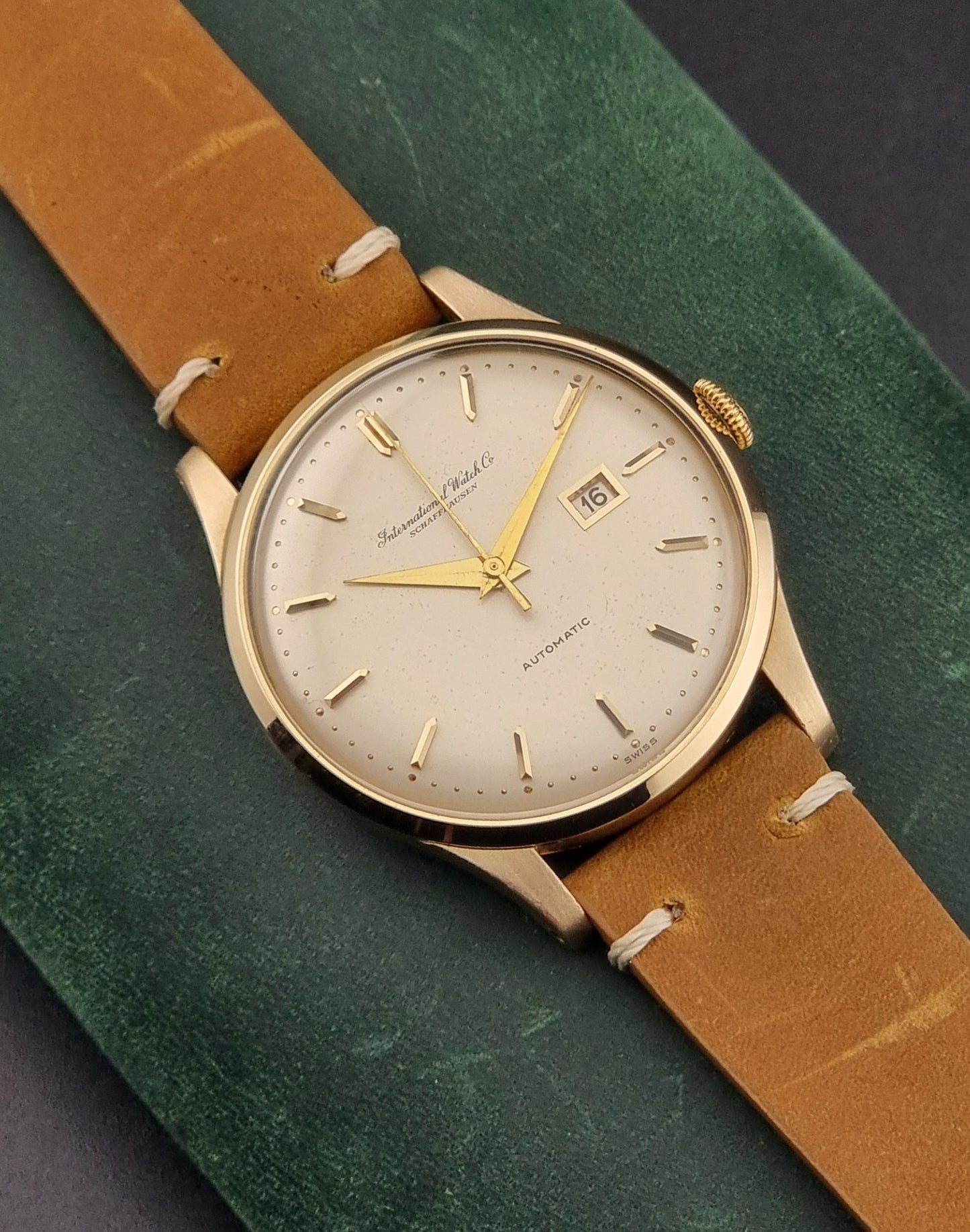 A vintage gold-plated IWC watch with a champagne dial, manual winding, and a brown leather strap, displayed against a green background.