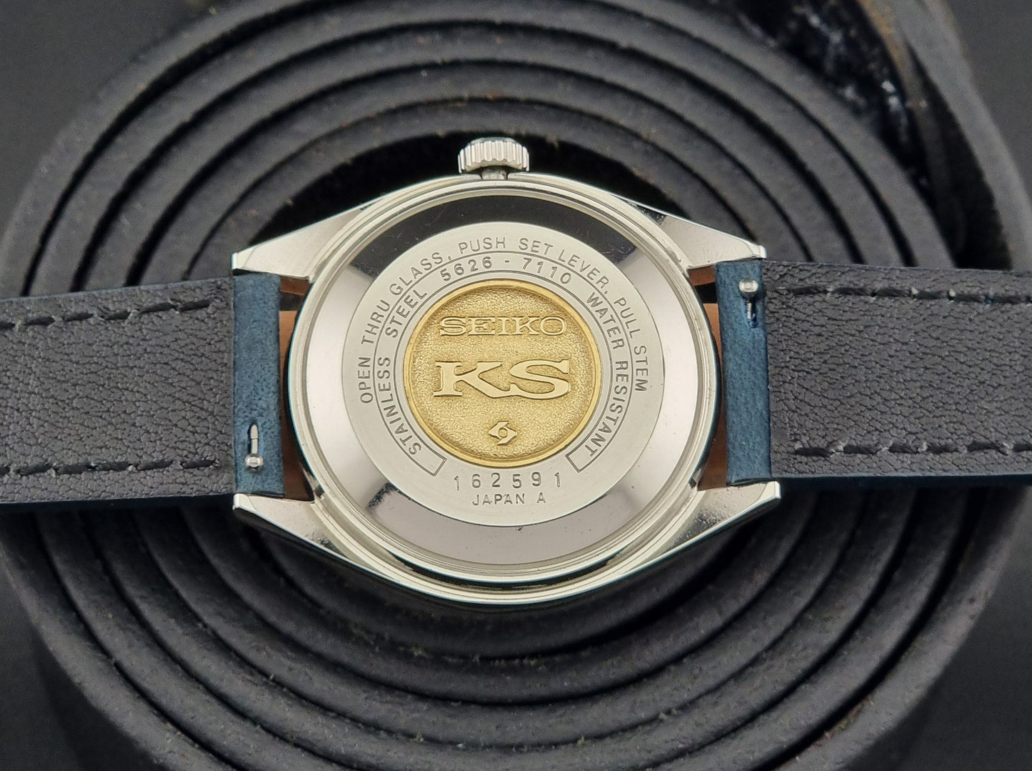 Vintage King Seiko 5626-7110 Wristwatch case back with blue strap and silver case on a dark textured background