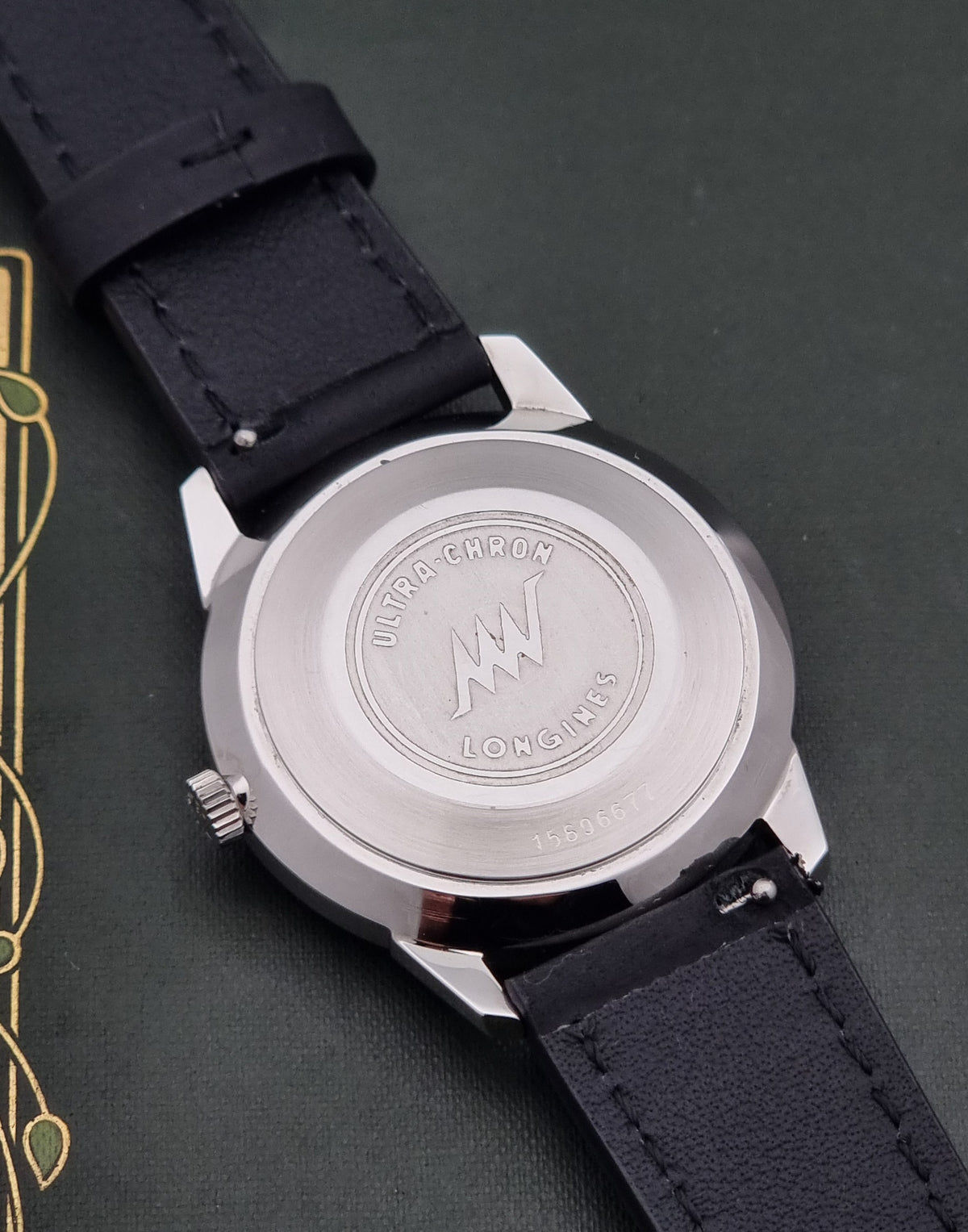 Close-up of the back of a Longines watch with a silver-toned case and black leather strap on a dark textured background.