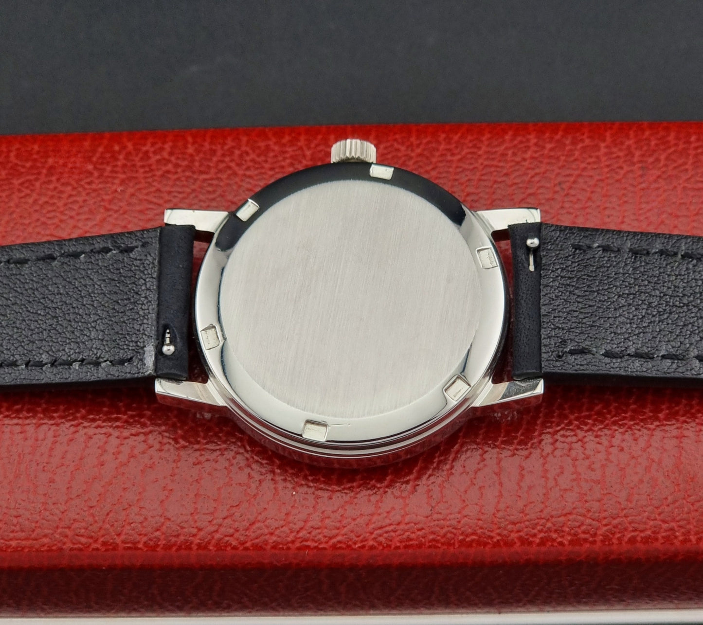Watch case back with a black leather strap on a red textured surface