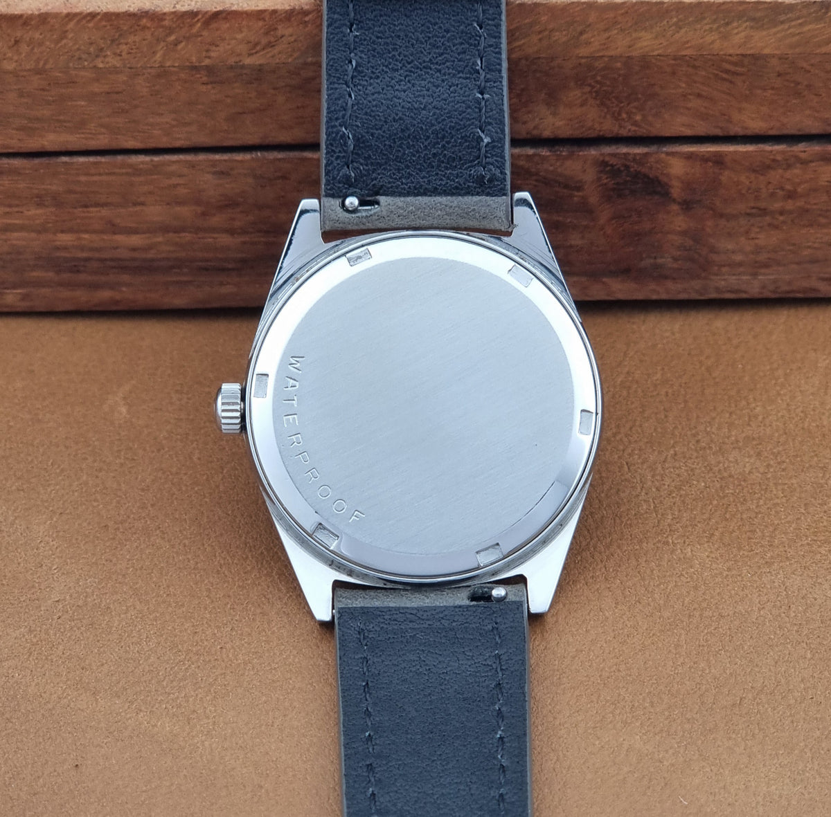 The back of a Vintage Omega Geneve 136.041 watch with a brushed stainless steel case and a dark blue leather strap, displayed on a brown leather surface with a wooden background.