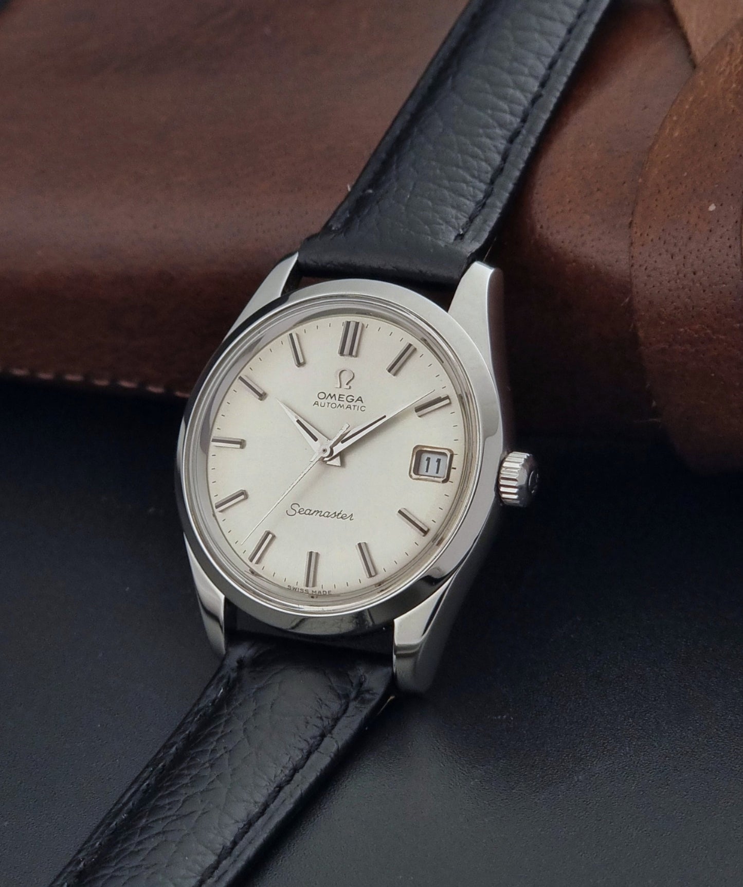 Vintage Omega Seamaster 166.010 watch with a silver dial and black leather strap on a dark surface crown view