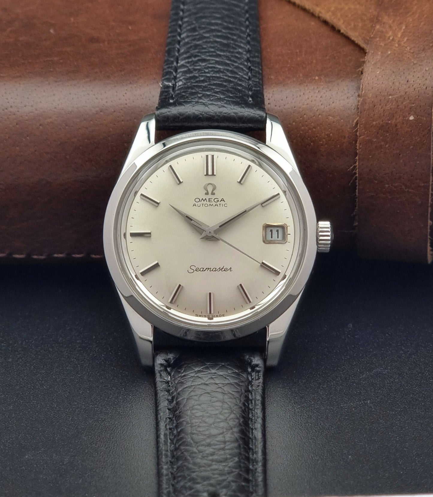 Vintage Omega Seamaster 166.010 watch with a silver dial and black leather strap on a dark surface front view