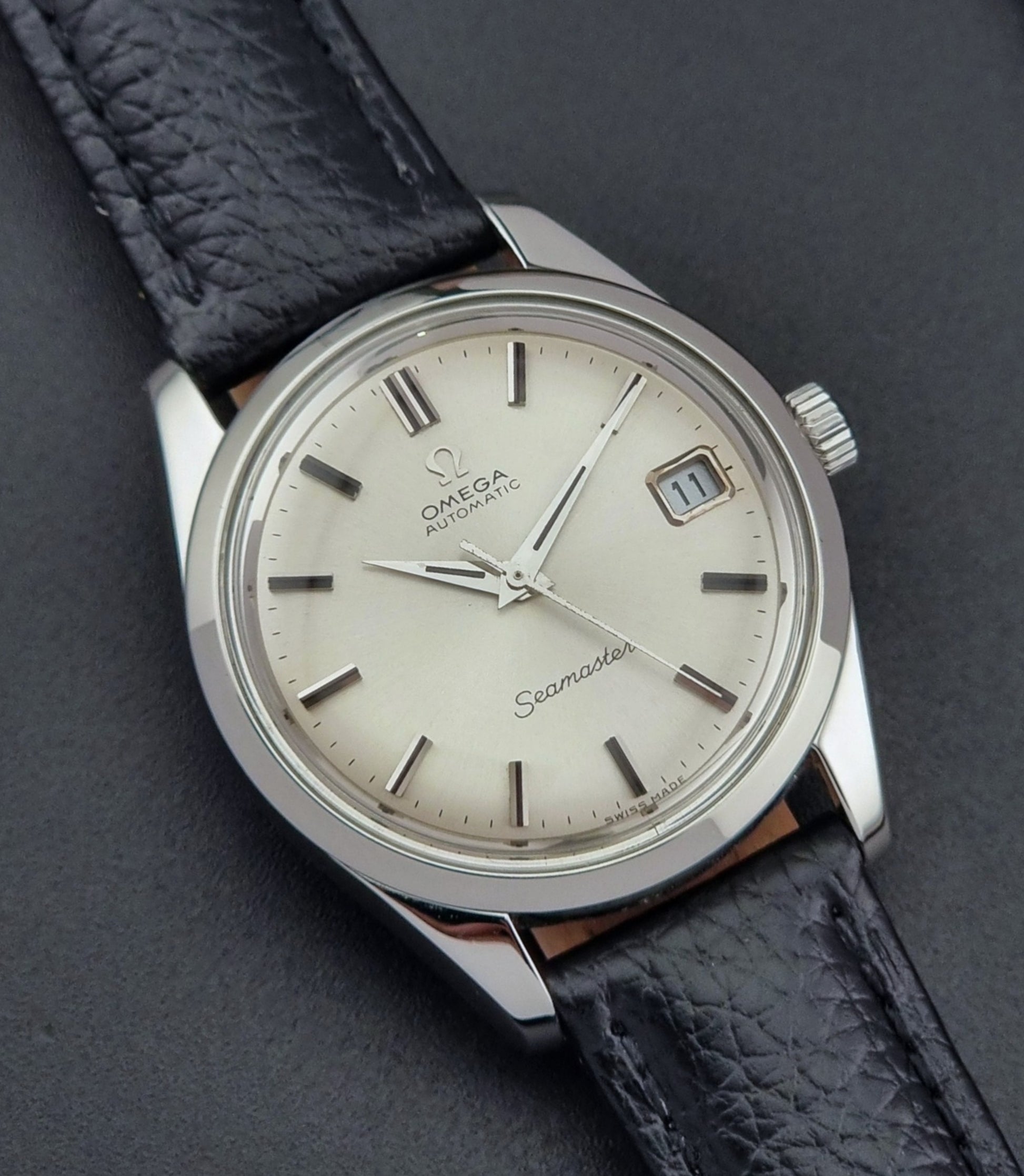 Vintage Omega Seamaster 166.010 watch with a silver dial and black leather strap on a dark surface close up dial