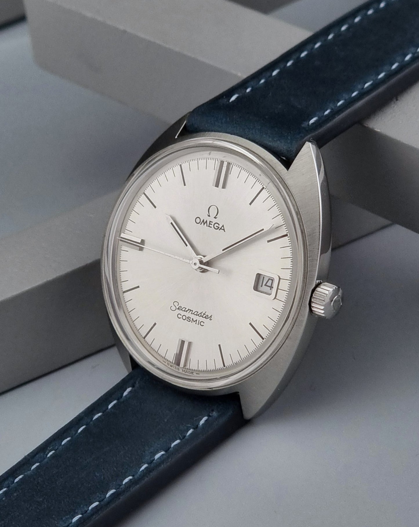 Vintage Omega Seamaster Cosmic wristwatch with blue strap and silver case on a gray background crown view