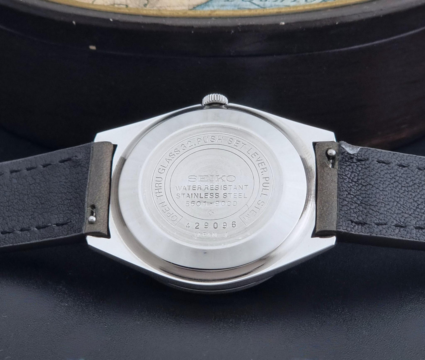 Close-up of the back of a Vintage Seiko Wristwatch LordMatic 5601-9000 with a silver stainless steel case and a dark gray leather strap, resting on a dark surface.