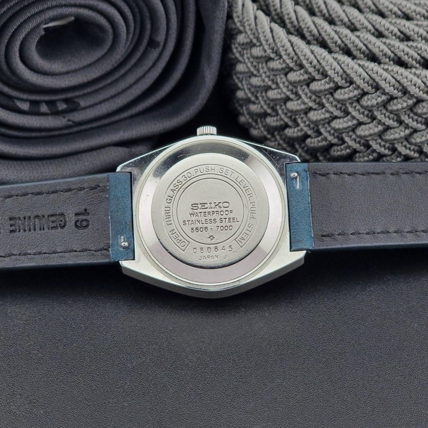 Vintage Seiko Wristwatch LordMatic 5606-7000 with blue strap on a textured gray background case back view