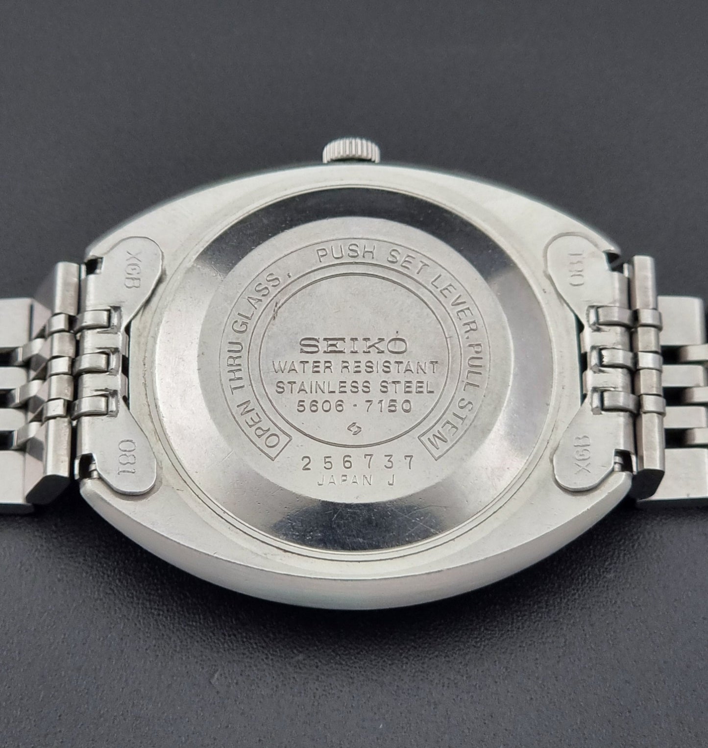 Vintage Seiko Lord Matic 5606-7150 stainless steel wristwatch with a metallic band on a dark gray background close up case back