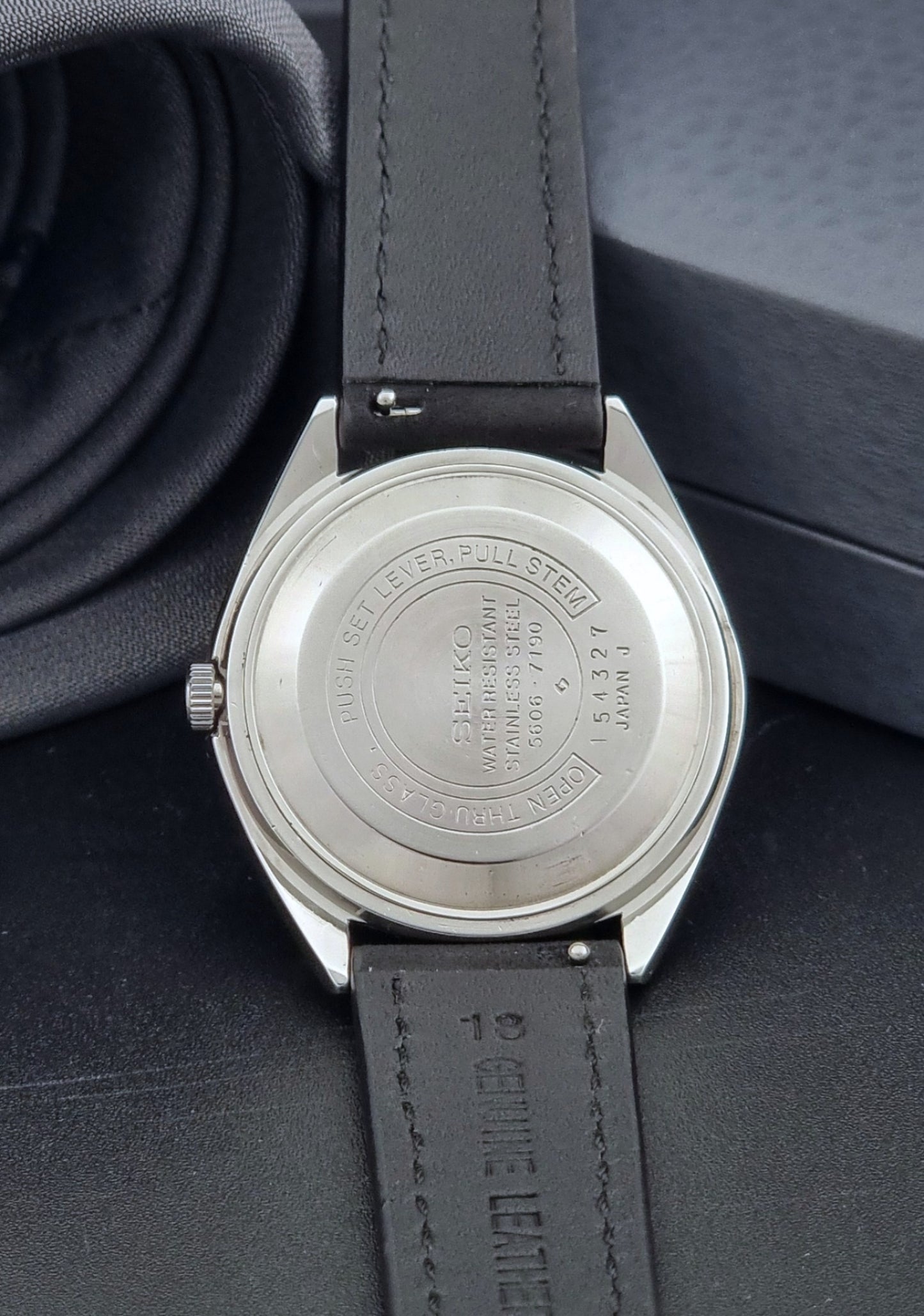 Vintage Seiko Wristwatch LordMatic 5606-7190 black strap and silver case on a dark gray background case back view
