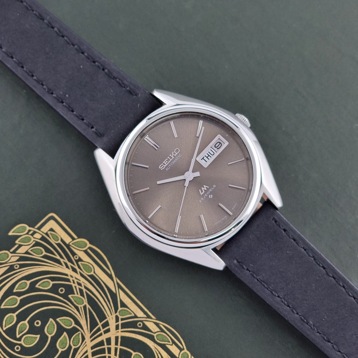 Vintage Seiko Lordmatic 5606-8031 watch with a brown dial and a dark strap on a green background