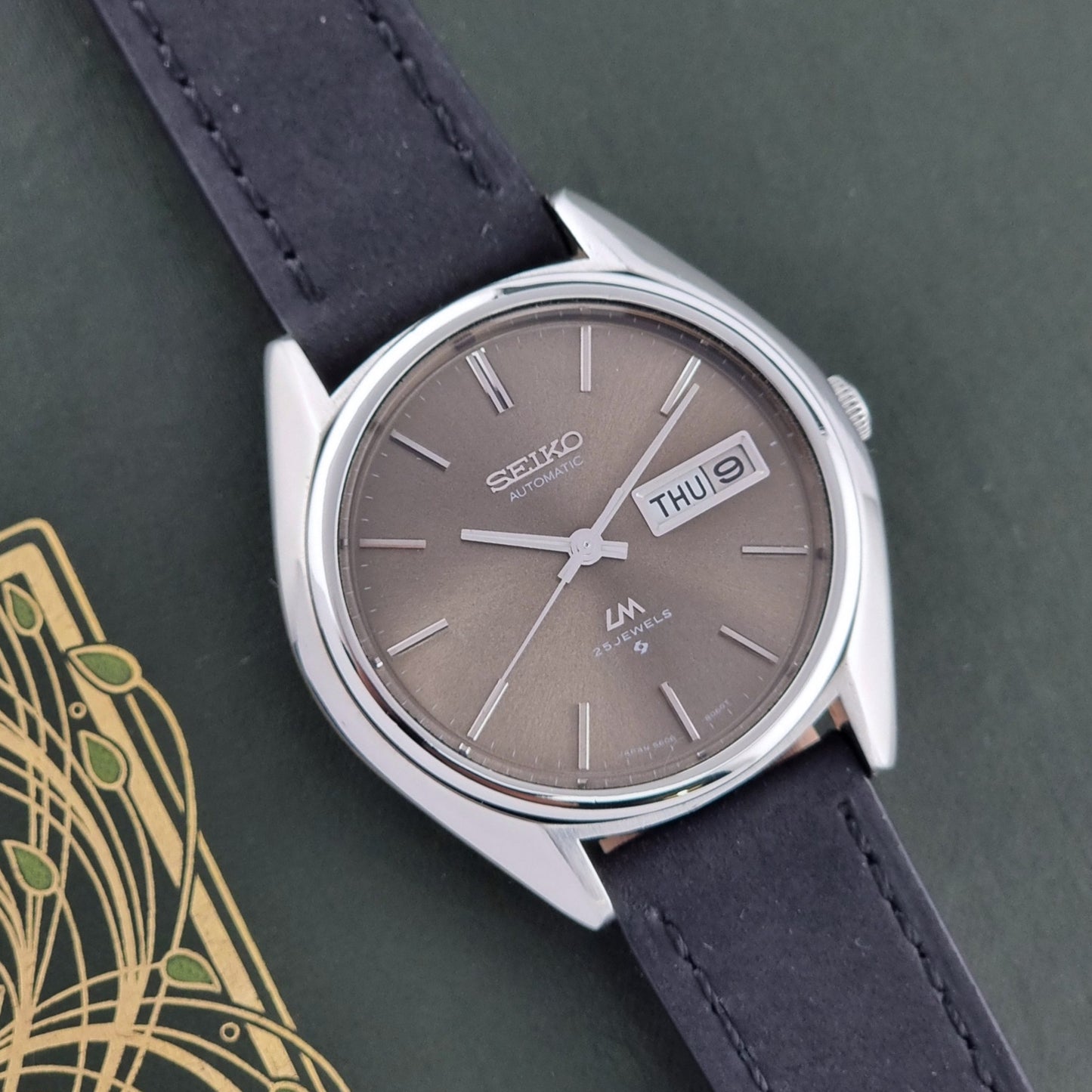 Vintage Seiko Lordmatic 5606-8031 watch with a brown dial and a dark strap on a green background