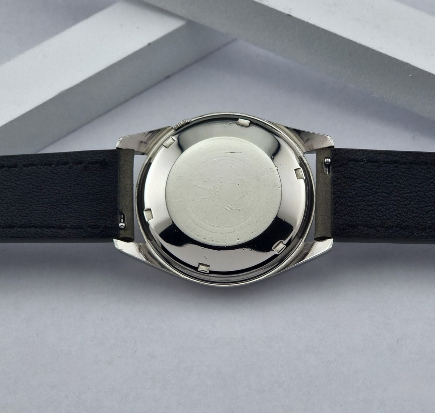 Vintage Seiko watch Seikomatic Weekdater 6218-8970 with a silver case and dark leather strap on a light gray background case back view