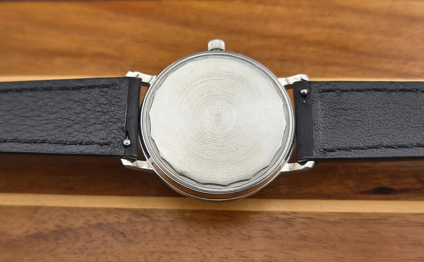Vintage Universal Geneve Watch case back with a black leather strap on a wooden surface