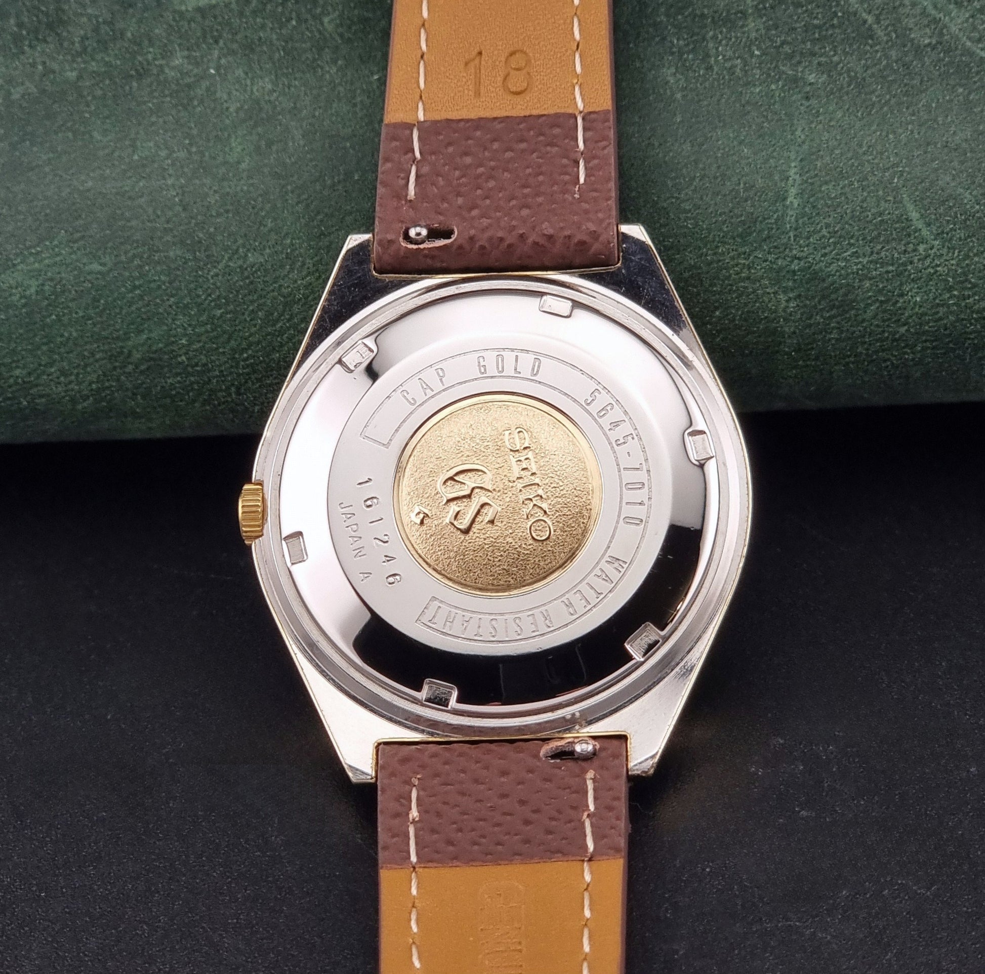 Vintage Grand Seiko Wristwatch 5645-7010 with a gold-tone case, cream dial, and brown textured leather strap, photographed on a dark gray background case back view