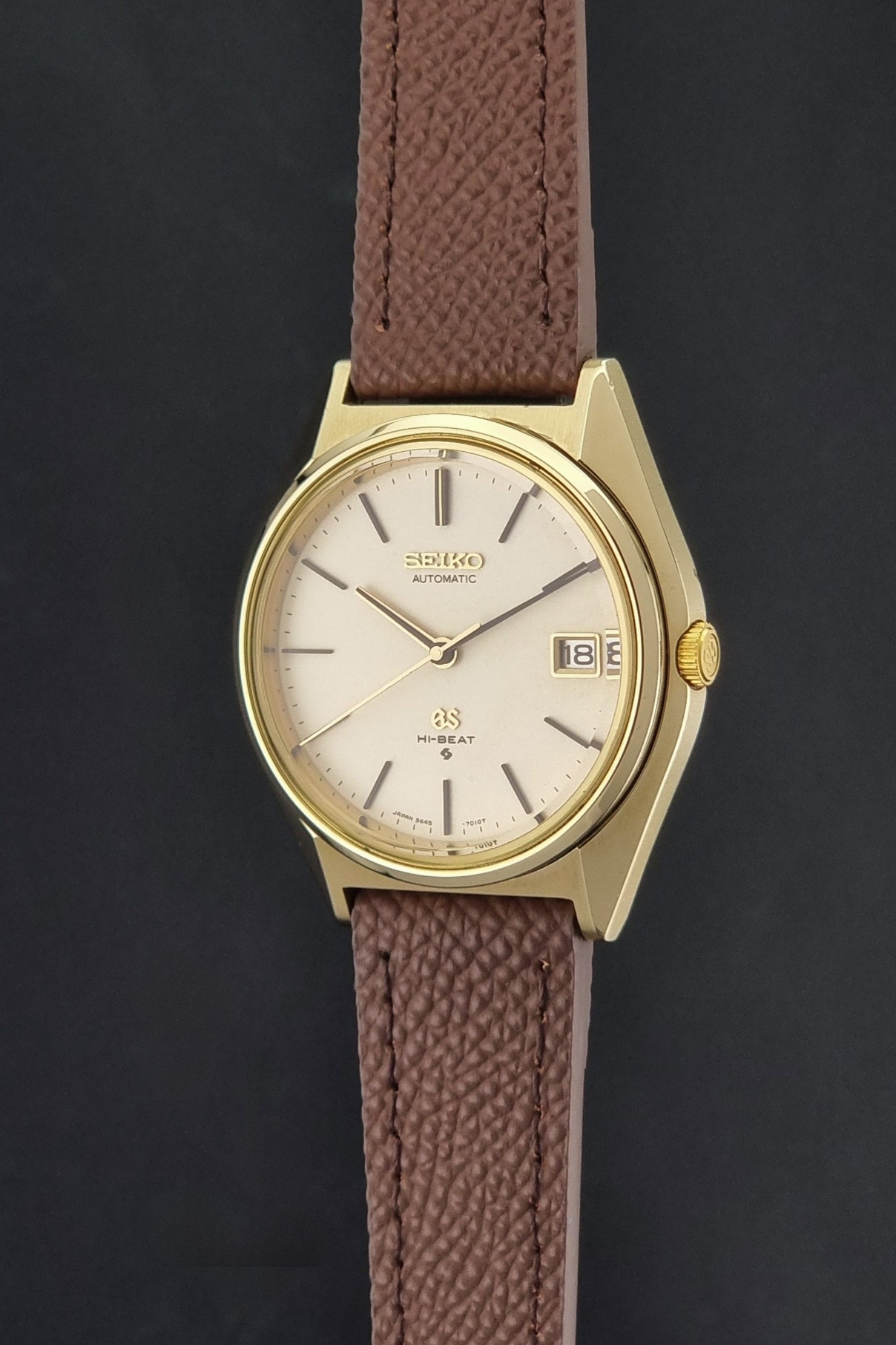 Vintage Grand Seiko Wristwatch 5645-7010 with a gold-tone case, cream dial, and brown textured leather strap, photographed on a dark gray background crown profile view