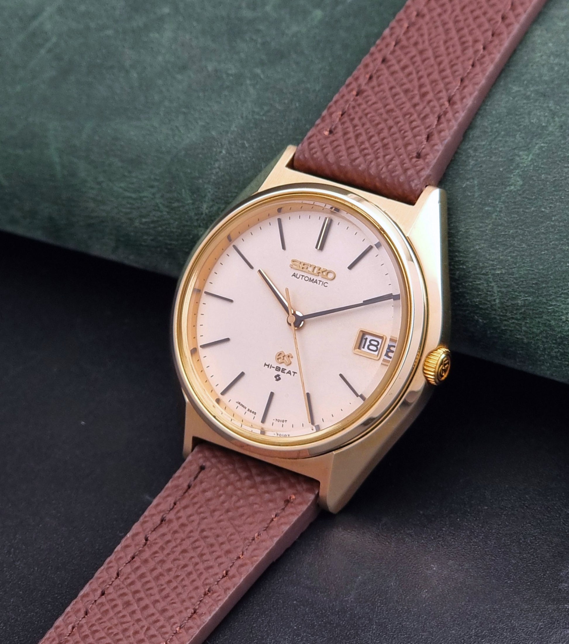 Vintage Grand Seiko Wristwatch 5645-7010 with a gold-tone case, cream dial, and brown textured leather strap, photographed on a dark gray background crown view