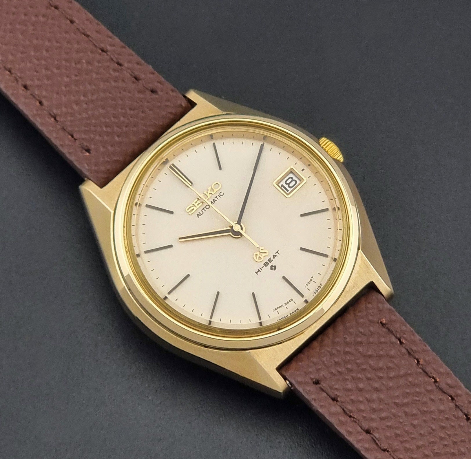 Vintage Grand Seiko Wristwatch 5645-7010 with a gold-tone case, cream dial, and brown textured leather strap, photographed on a dark gray background close dial view