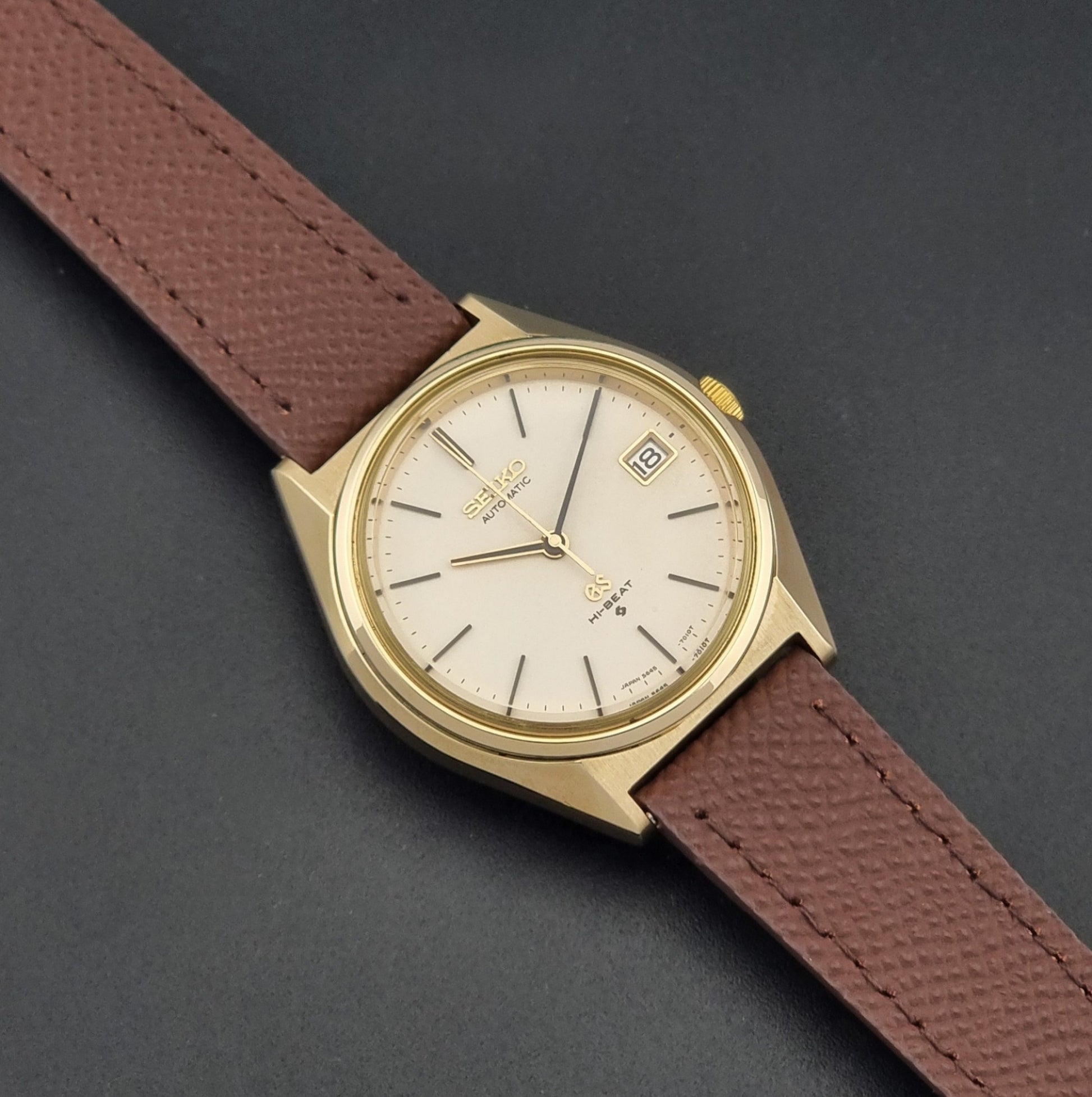 Vintage Grand Seiko Wristwatch 5645-7010 with a gold-tone case, cream dial, and brown textured leather strap, photographed on a dark gray background dial
