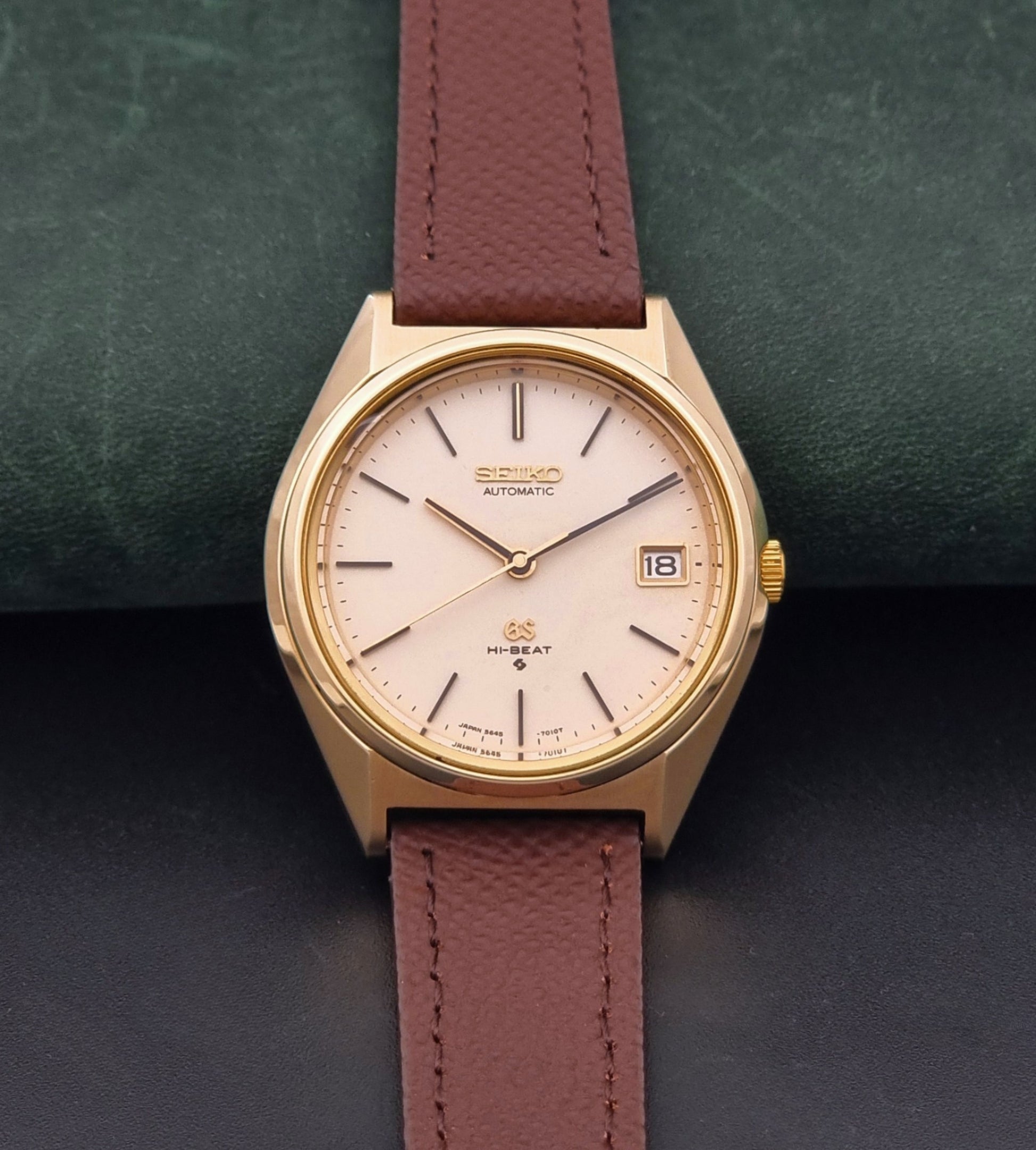 Vintage Grand Seiko Wristwatch 5645-7010 with a gold-tone case, cream dial, and brown textured leather strap, photographed on a dark gray background dial view