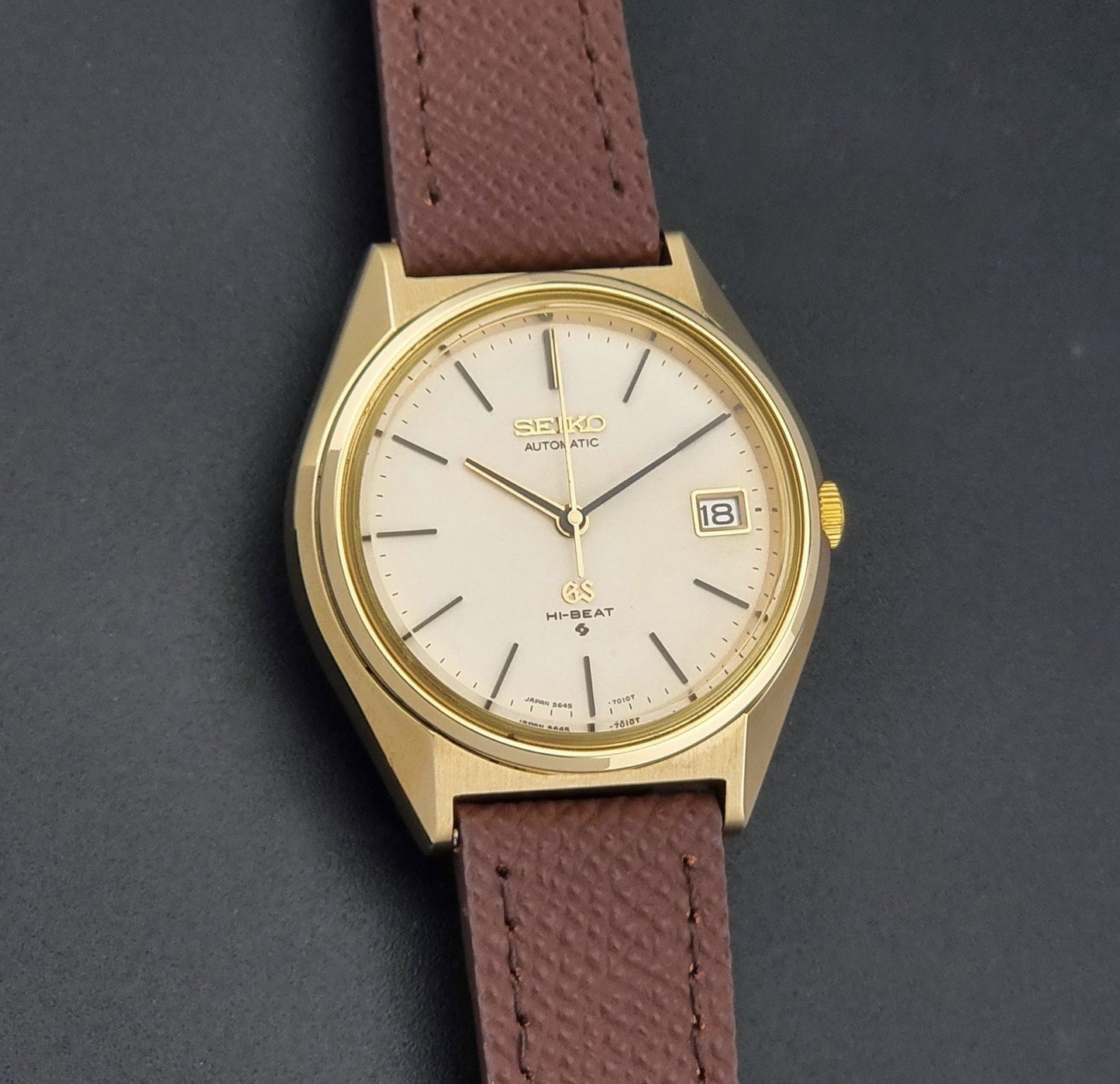 Vintage Grand Seiko Wristwatch 5645-7010 with a gold-tone case, cream dial, and brown textured leather strap, photographed on a dark gray background front view