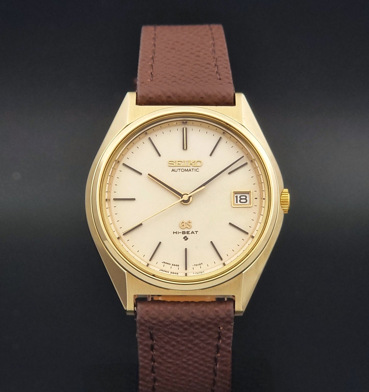 Vintage Grand Seiko Wristwatch 5645-7010 with a gold-tone case, cream dial, and brown textured leather strap, photographed on a dark gray background dial view