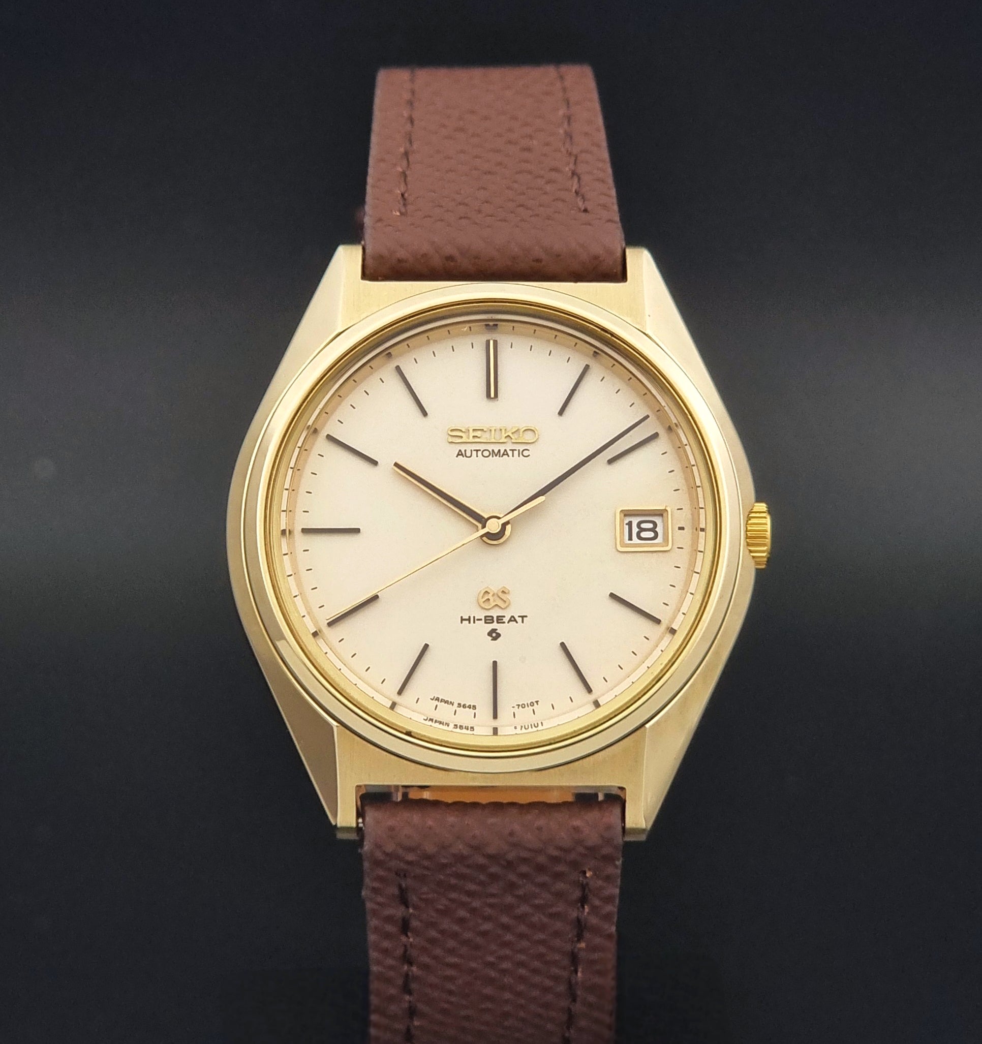 Vintage Grand Seiko Wristwatch 5645-7010 with a gold-tone case, cream dial, and brown textured leather strap, photographed on a dark gray background dial view