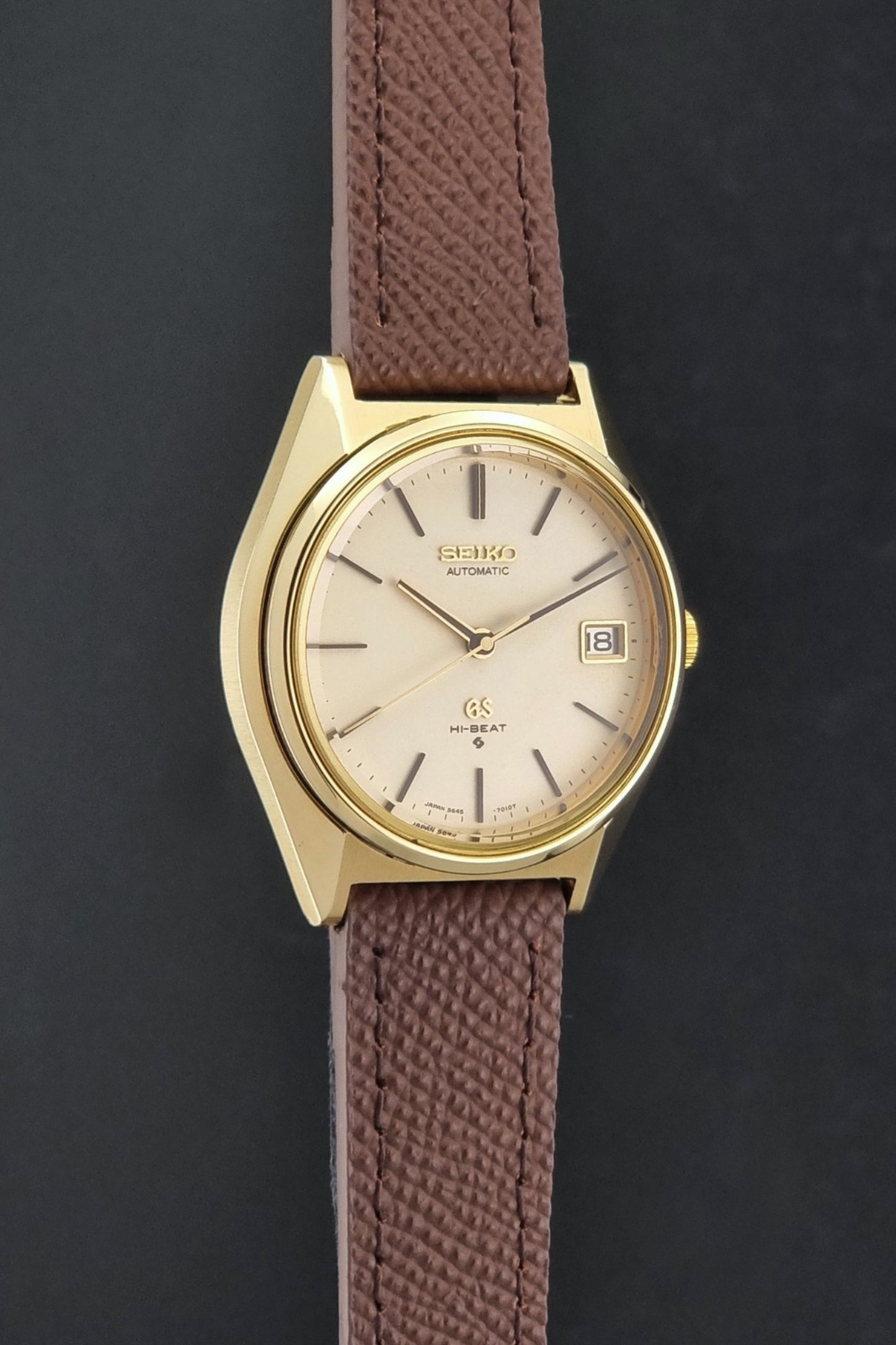 Vintage Grand Seiko Wristwatch 5645-7010 with a gold-tone case, cream dial, and brown textured leather strap, photographed on a dark gray background side profile view