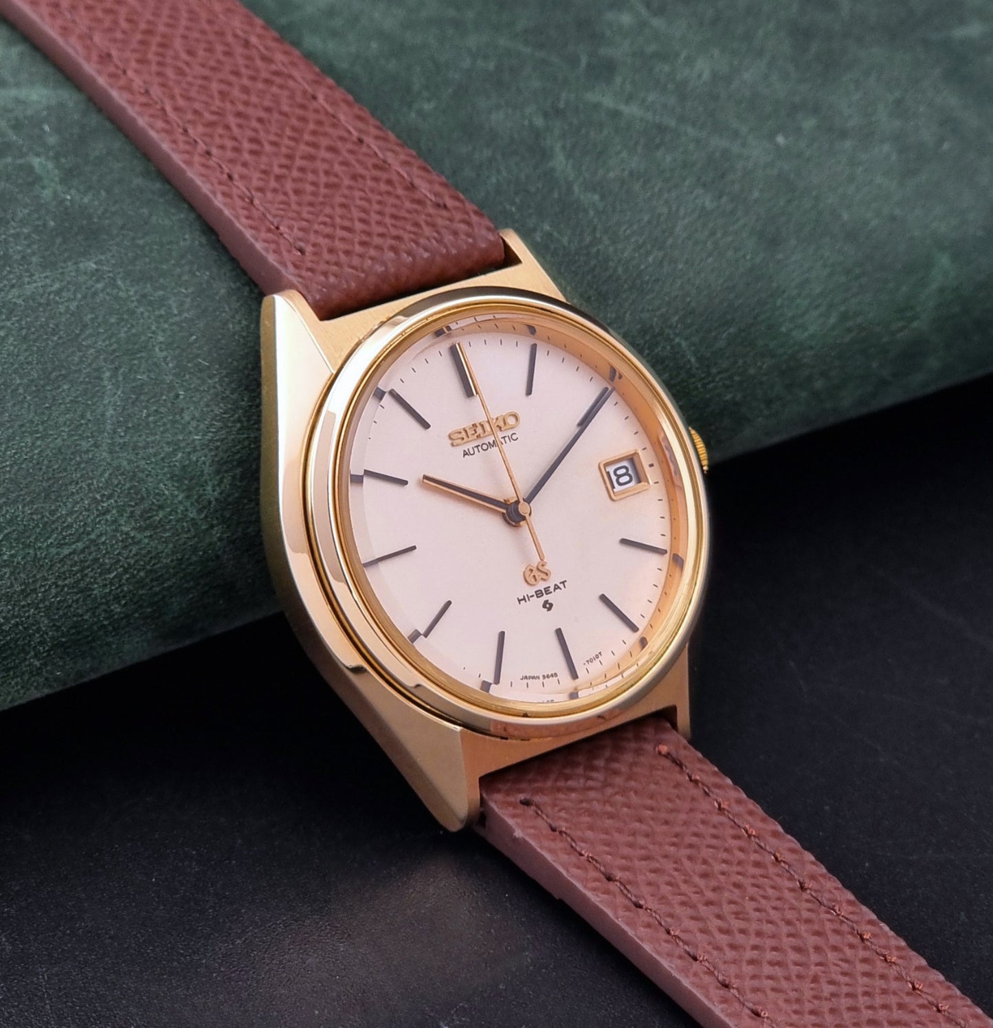 Vintage Grand Seiko Wristwatch 5645-7010 with a gold-tone case, cream dial, and brown textured leather strap, photographed on a dark gray background dial view
