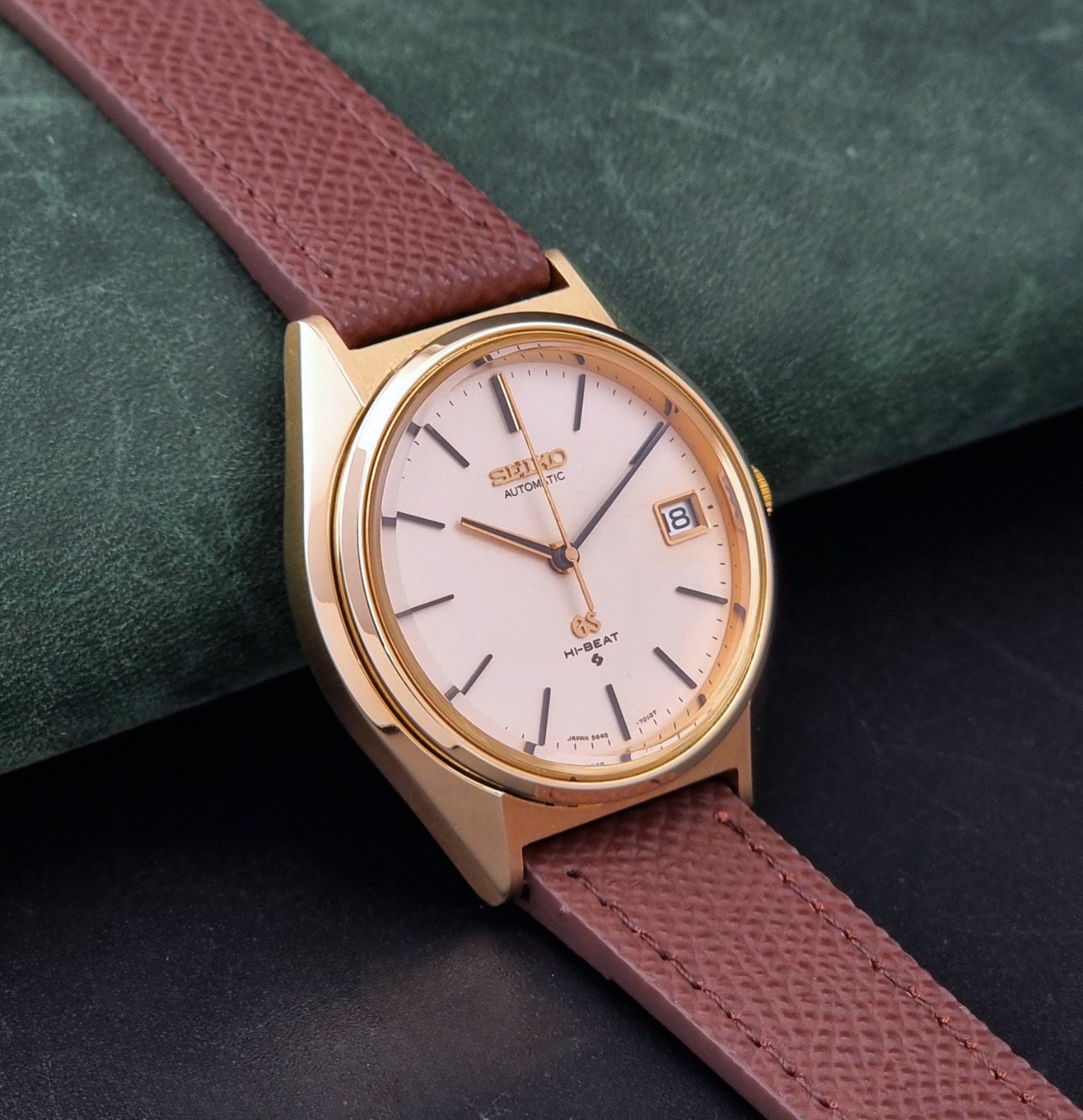 Vintage Grand Seiko Wristwatch 5645-7010 with a gold-tone case, cream dial, and brown textured leather strap, photographed on a dark gray background dial view