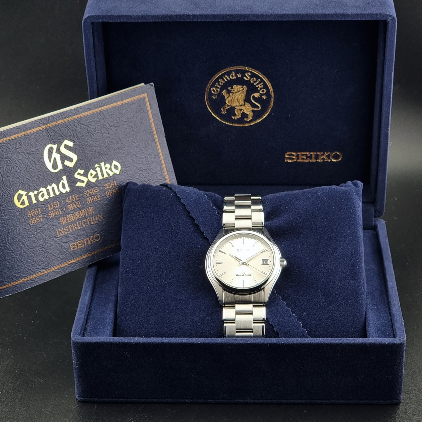 A Grand Seiko stainless steel wristwatch with a silver link bracelet and white dial, presented inside an open dark blue velvet watch box with branded insert and a booklet.