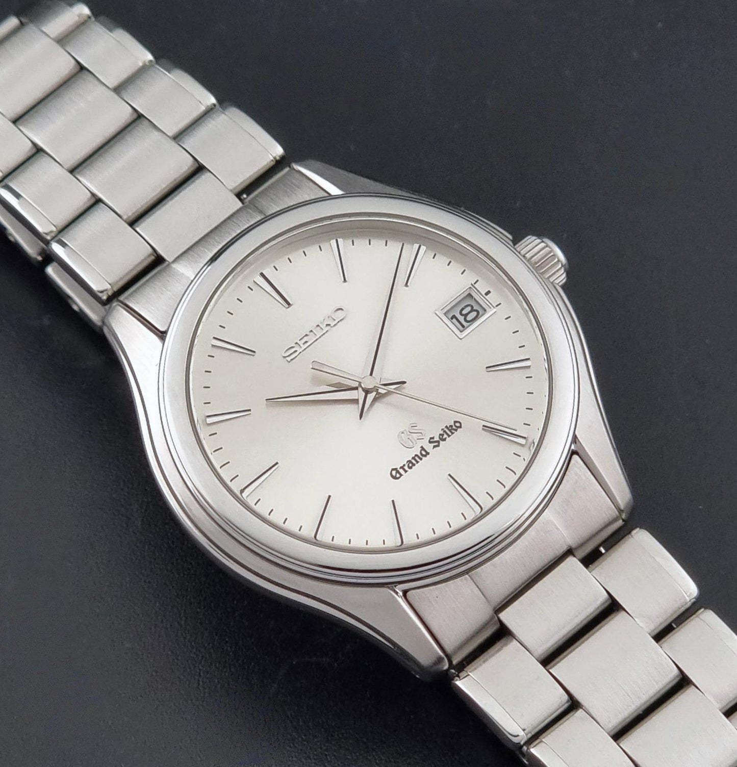 A Grand Seiko SBGX005 quartz watch 9F62-0A10 Sunburst Dial with bracelet on a dark back ground with a silver-tone metal link bracelet, and a date window, shown on a dark textured background.