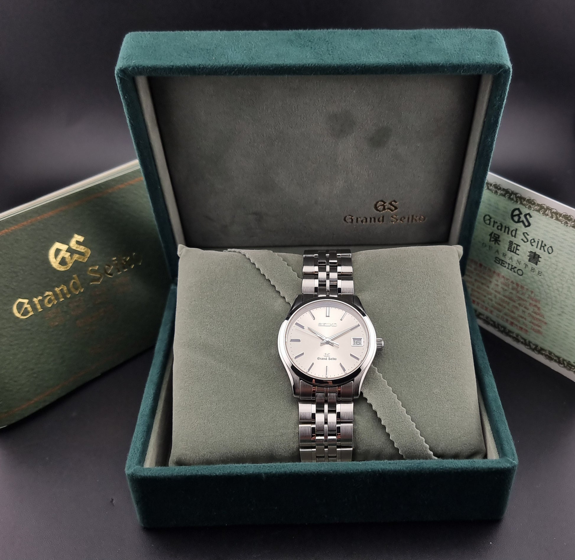 A Grand Seiko SBGV001 quartz watch and link bracelet and a cream-colored dial, presented inside an open dark green velvet watch box with a matching watch certificate and box lid visible.
