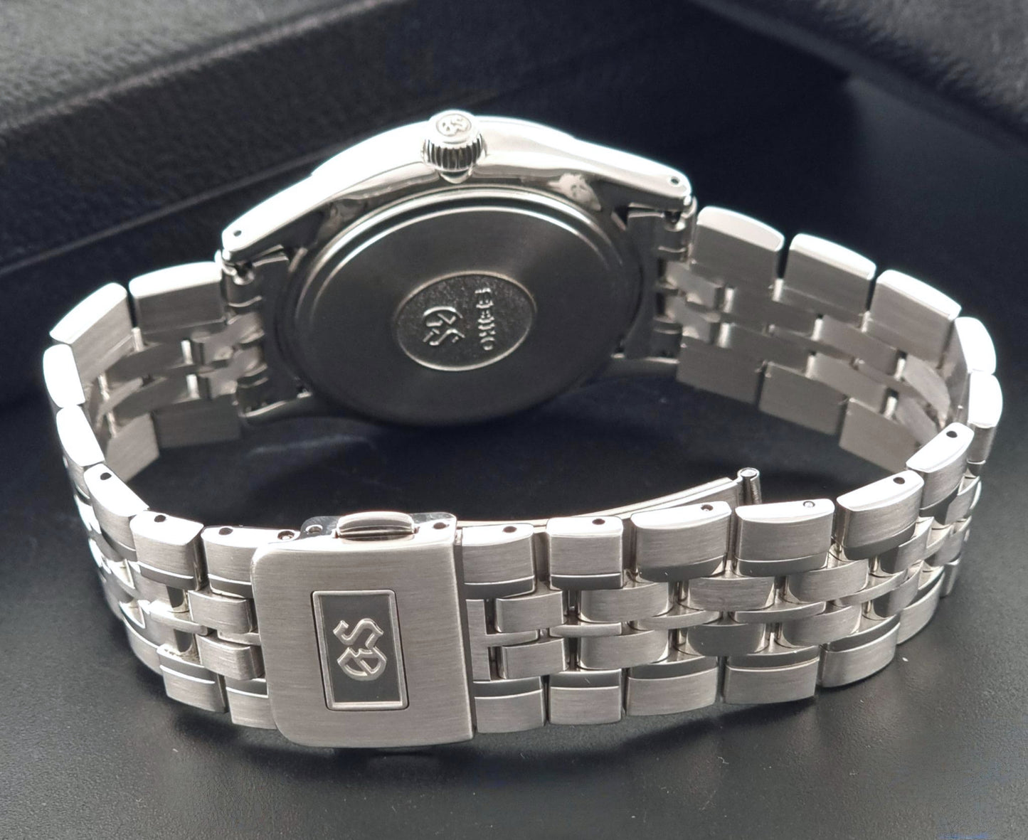 A Grand Seiko SBGV001 quartz watch and link bracelet, shown from the back, featuring a brushed metal clasp with a logo and a circular case back plate with model markings, photographed on a dark textured surface.