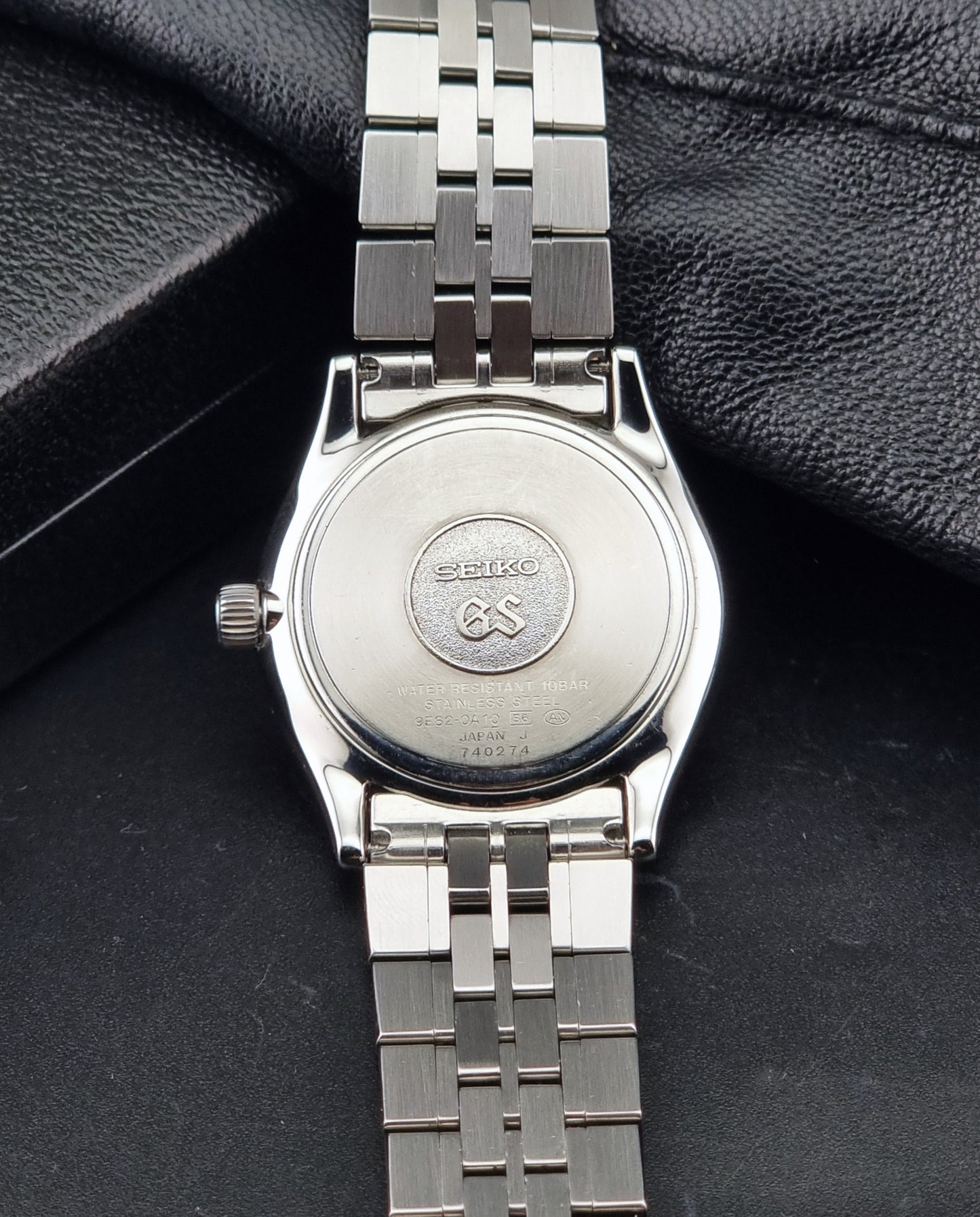 The back of A Grand Seiko SBGV001 quartz watch and link bracelet, resting on a textured black surface. The brand name 'SEIKO GS' is engraved on the watch case.