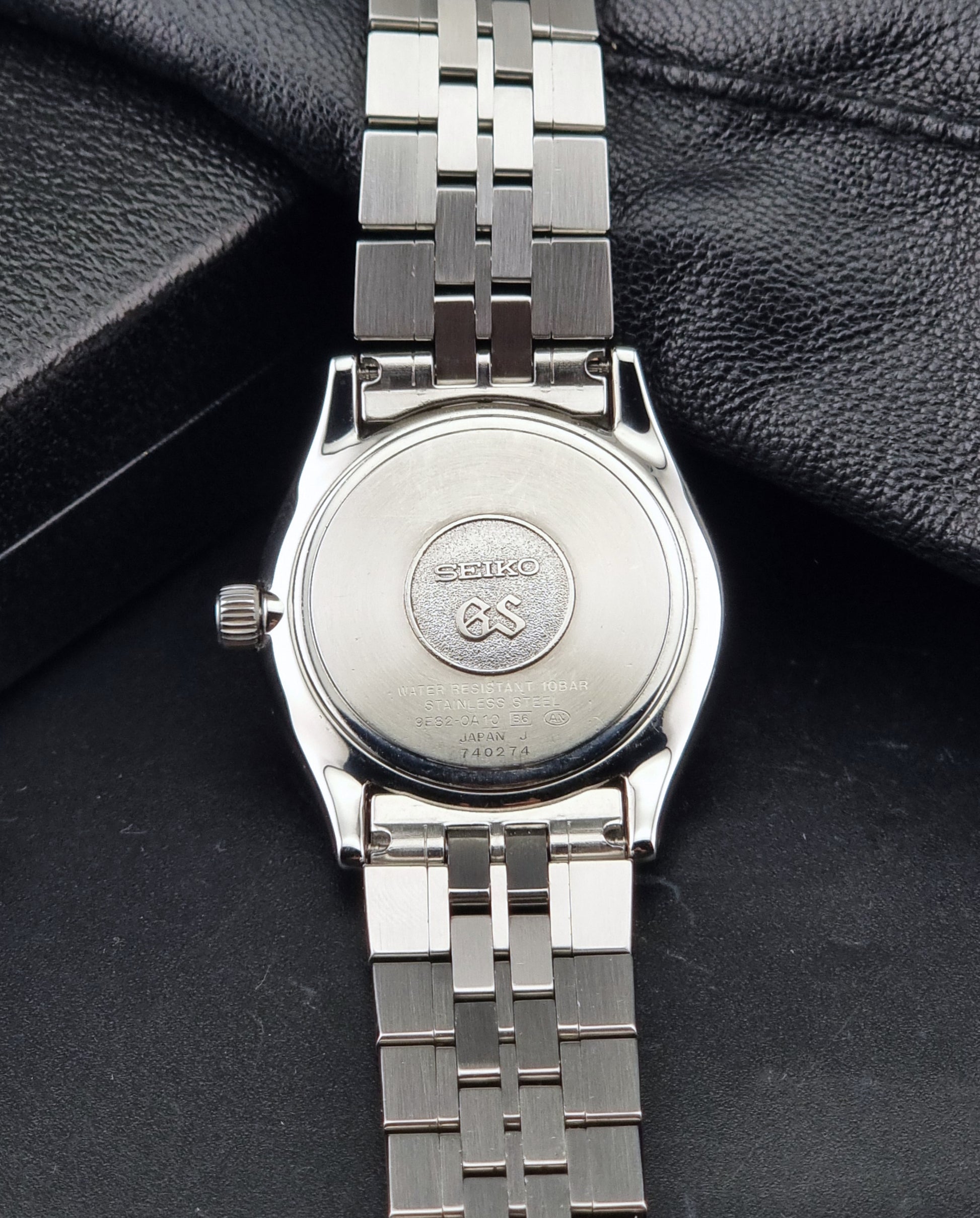 The back of A Grand Seiko SBGV001 quartz watch and link bracelet, resting on a textured black surface. The brand name 'SEIKO GS' is engraved on the watch case.