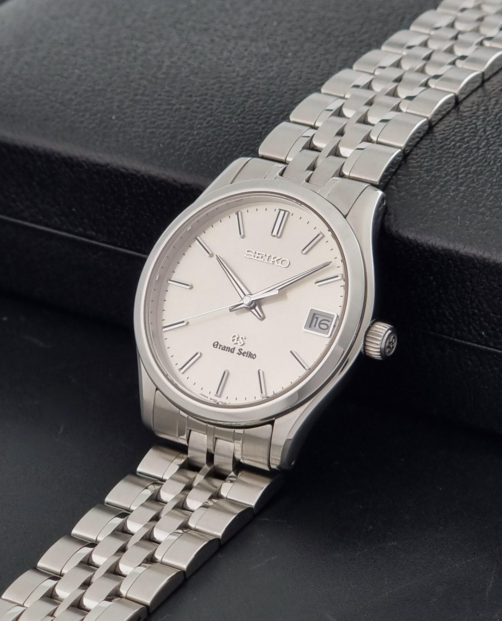 A Grand Seiko SBGV001 quartz watch and link bracelet and a cream-colored dial, resting on a textured black surface.