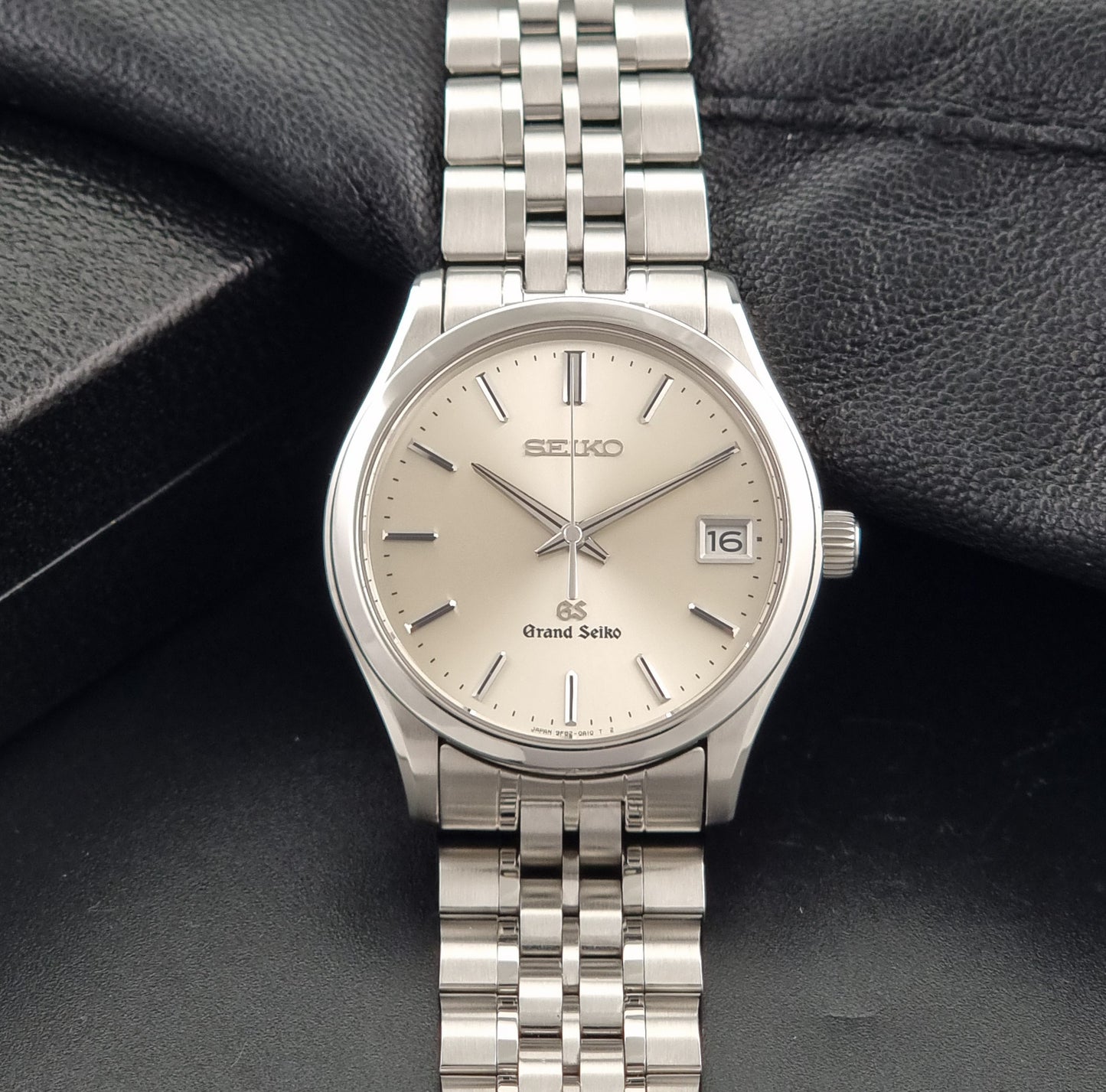 A Grand Seiko SBGV001 quartz watch and link bracelet with a champagne dial, resting on a textured black leather surface.