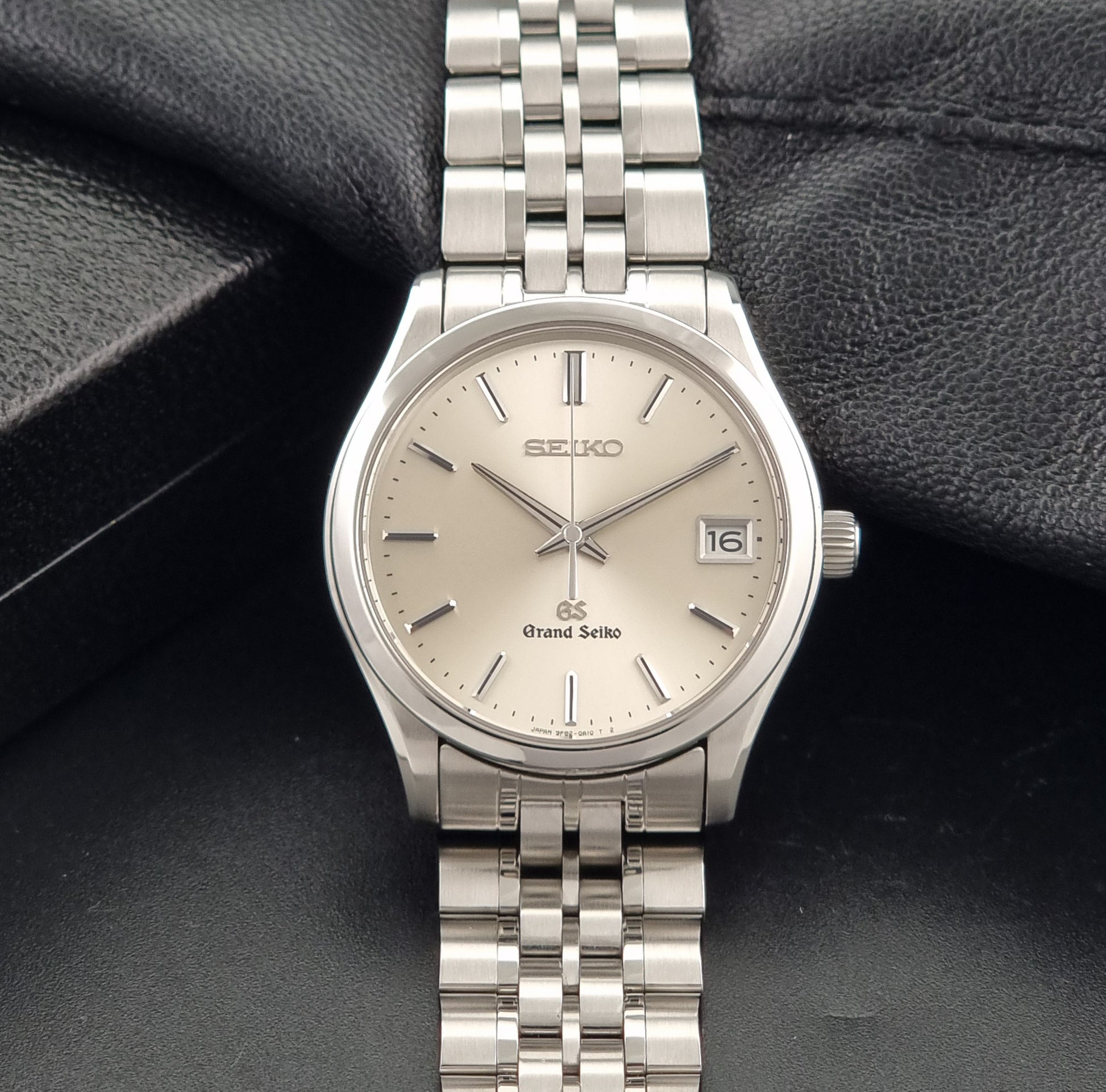 A Grand Seiko SBGV001 quartz watch and link bracelet with a champagne dial, resting on a textured black leather surface.