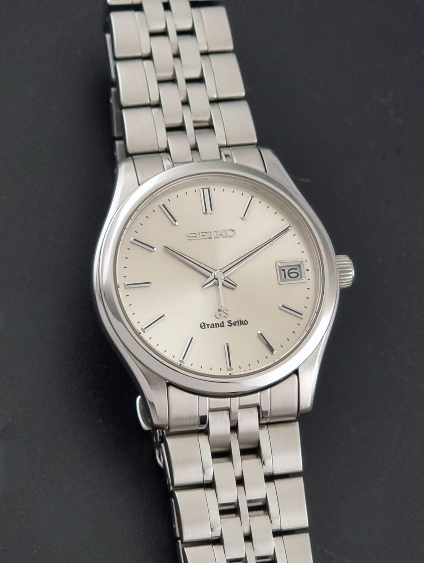 A Grand Seiko SBGV001 quartz watch and link bracelet and a cream-colored dial, shown on a dark gray background.
