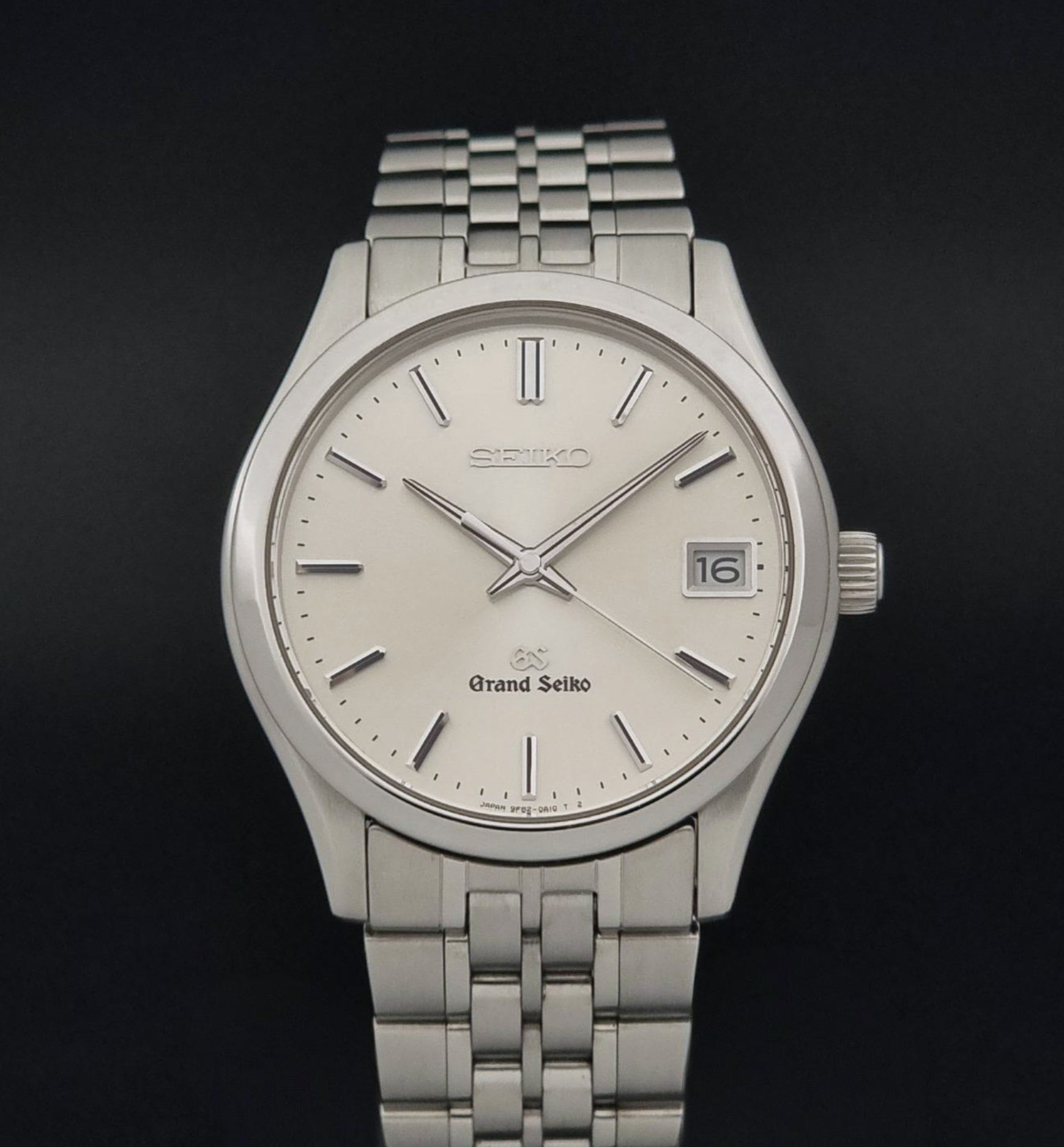 A Grand Seiko SBGV001 quartz watch and link bracelet with a cream-colored dial, and a date window, shown against a dark background.