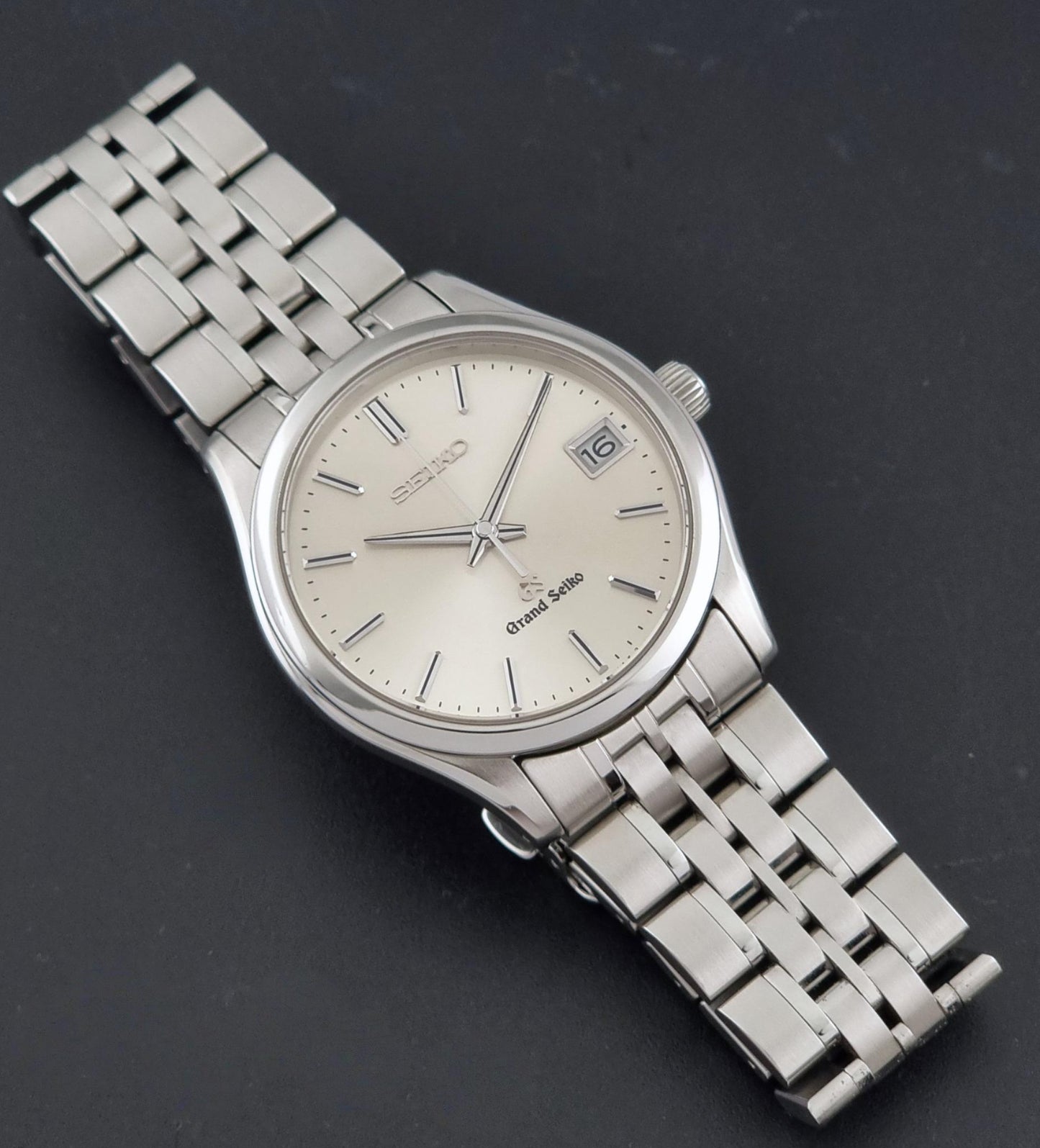 A Grand Seiko SBGV001 quartz watch and link bracelet, featuring a cream-colored dial with silver indices and a date window at the 3 o'clock position, shown on a dark gray background.