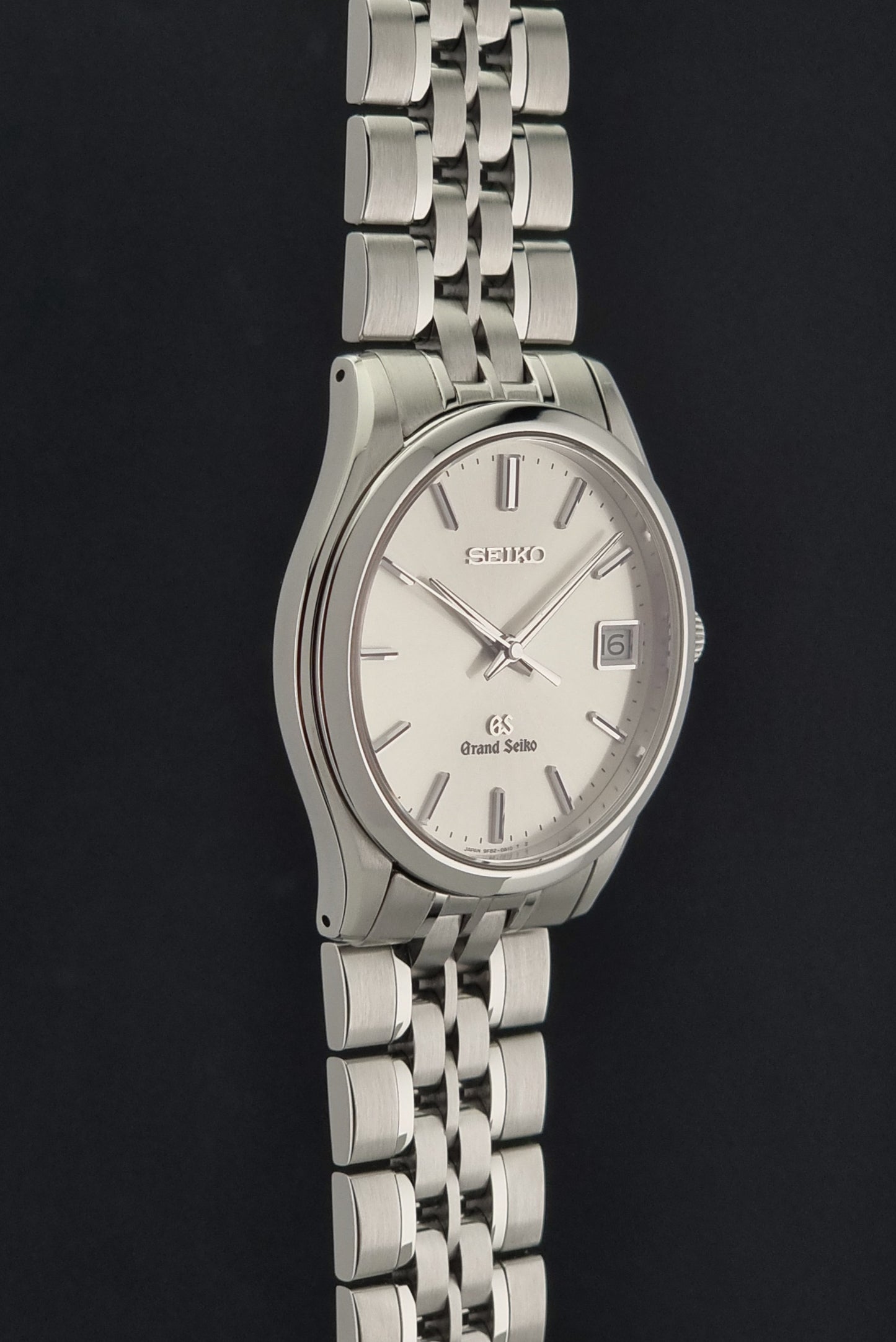 A Grand Seiko SBGV001 quartz watch and link bracelet and a cream-colored dial, with silver-tone hour markers and a date window at 3 o'clock, set against a dark background.